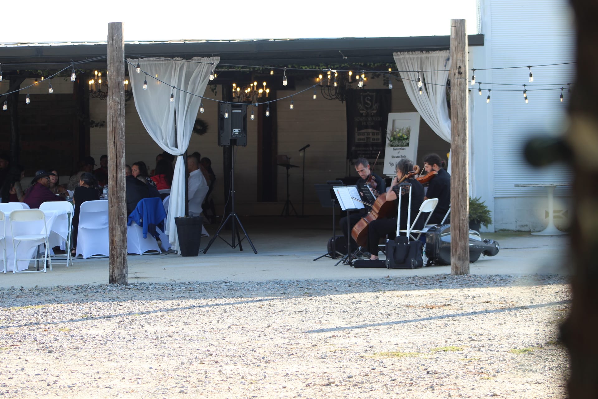 A string trio performs for wedding guests seated at draped tables under warm bistro lights at Scarborough House venue.