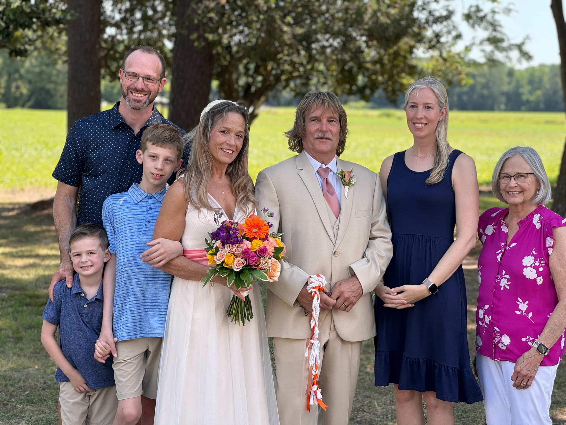Bride in white gown holding a colorful bouquet stands beside groom in a tan suit surrounded by smiling family members including two young boys against a backdrop of oak trees and green fields at Scarborough House.