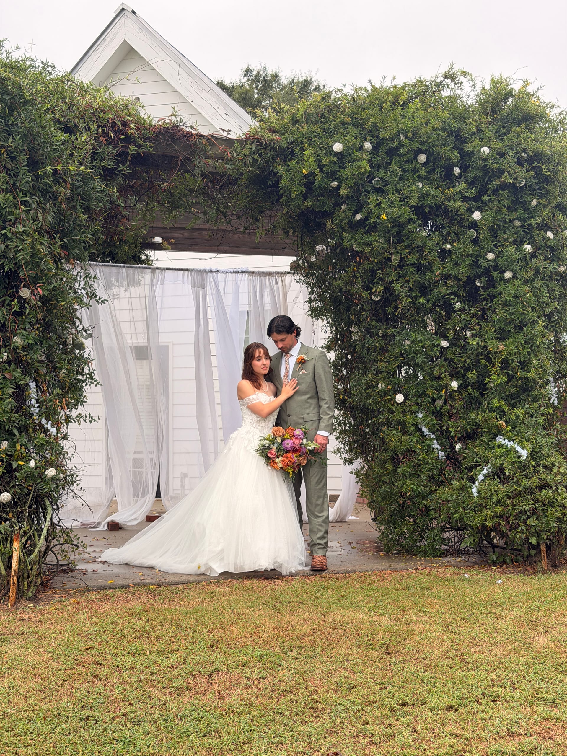 Bride in off-shoulder white gown holding a colorful bouquet leans toward groom in sage suit beneath a verdant ivy arch with white draping at Scarborough House historic estate.