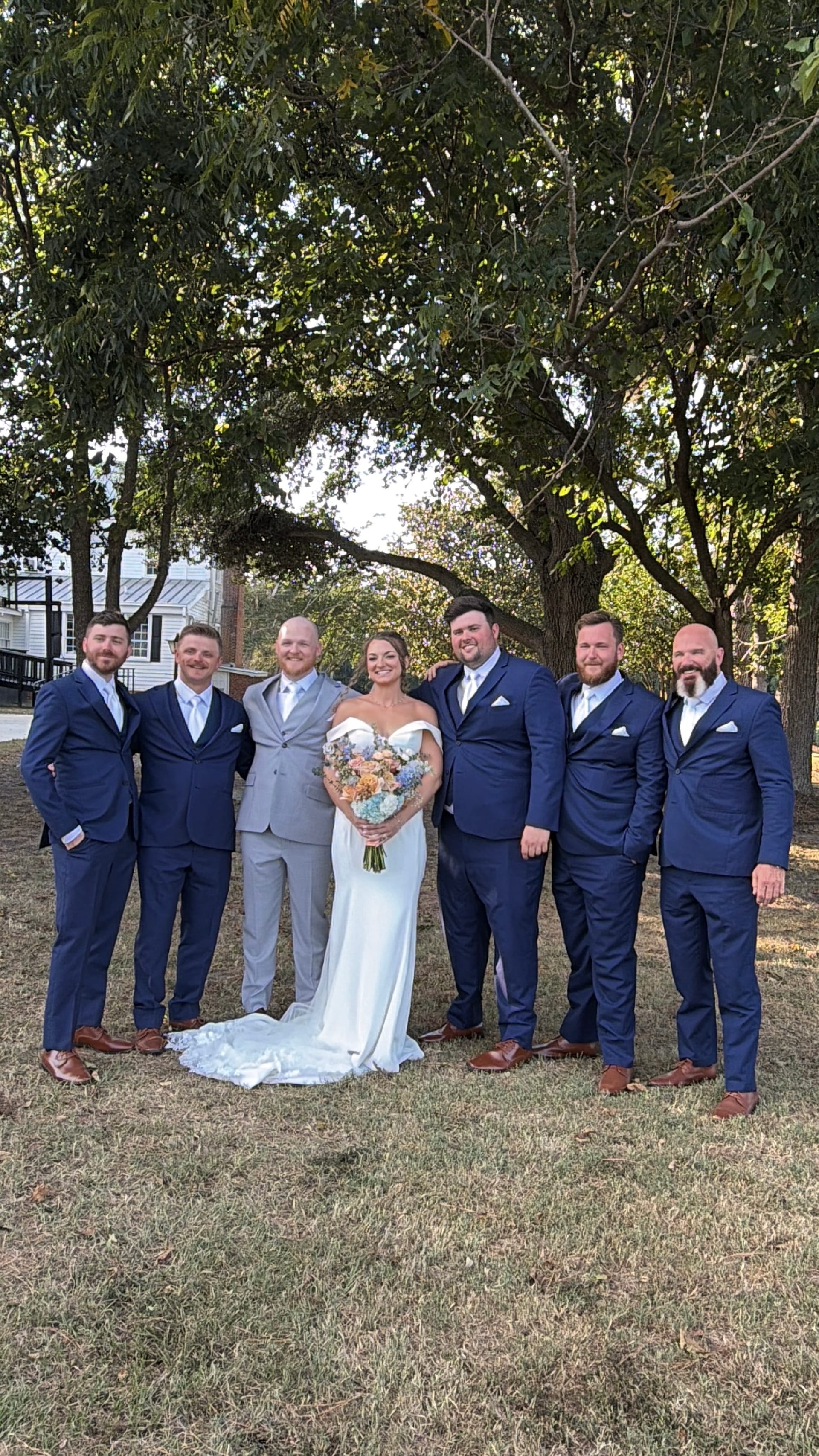 Bride in a white off-shoulder gown holding a pastel bouquet poses with five groomsmen in navy suits and the groom in a grey suit beneath large oak trees on the ceremony lawn at Scarborough House.