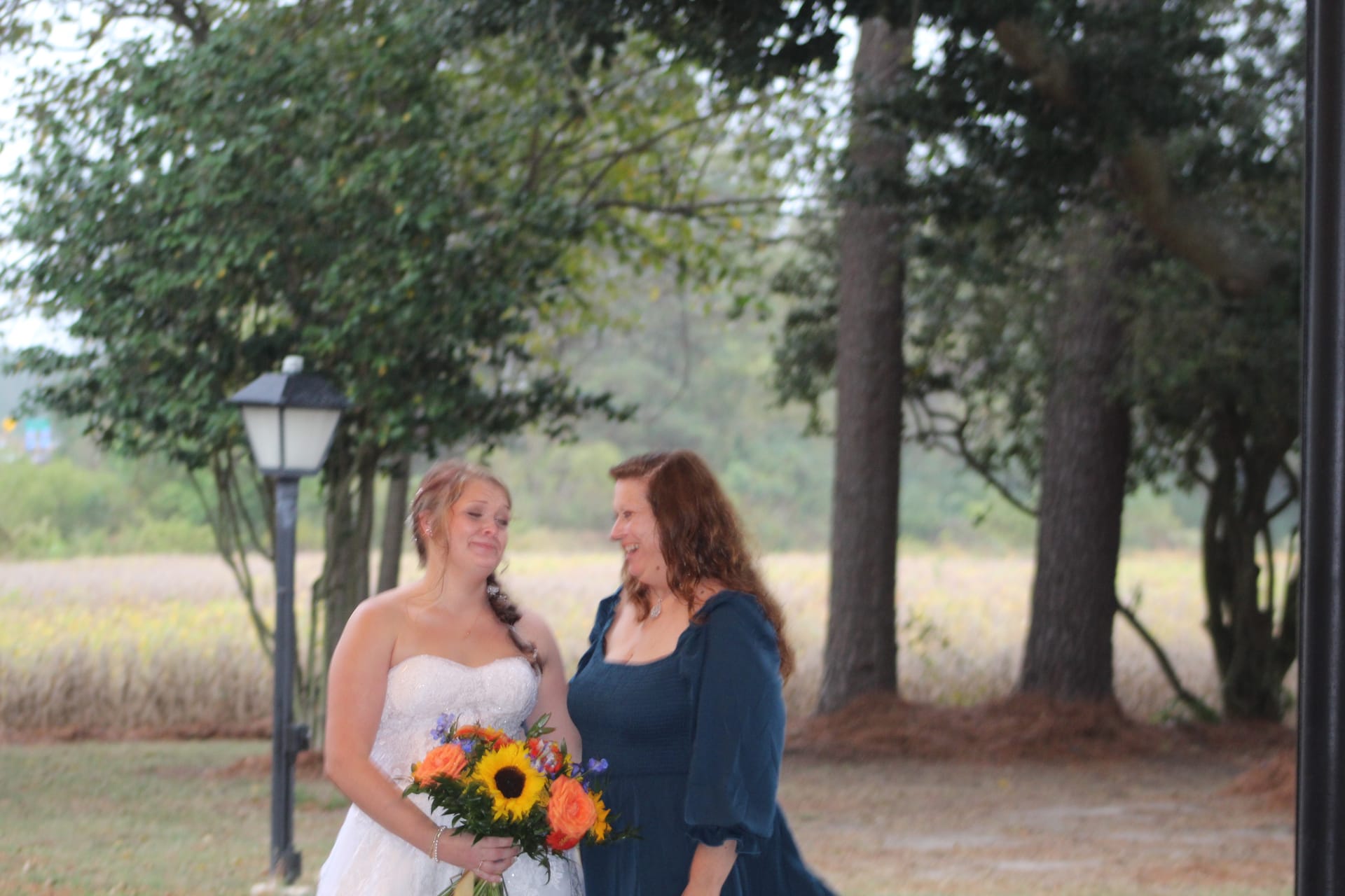Bride in a strapless white gown holding a sunflower and orange rose bouquet shares a warm smile with a woman in a navy dress against a scenic farmland backdrop at Scarborough House.
