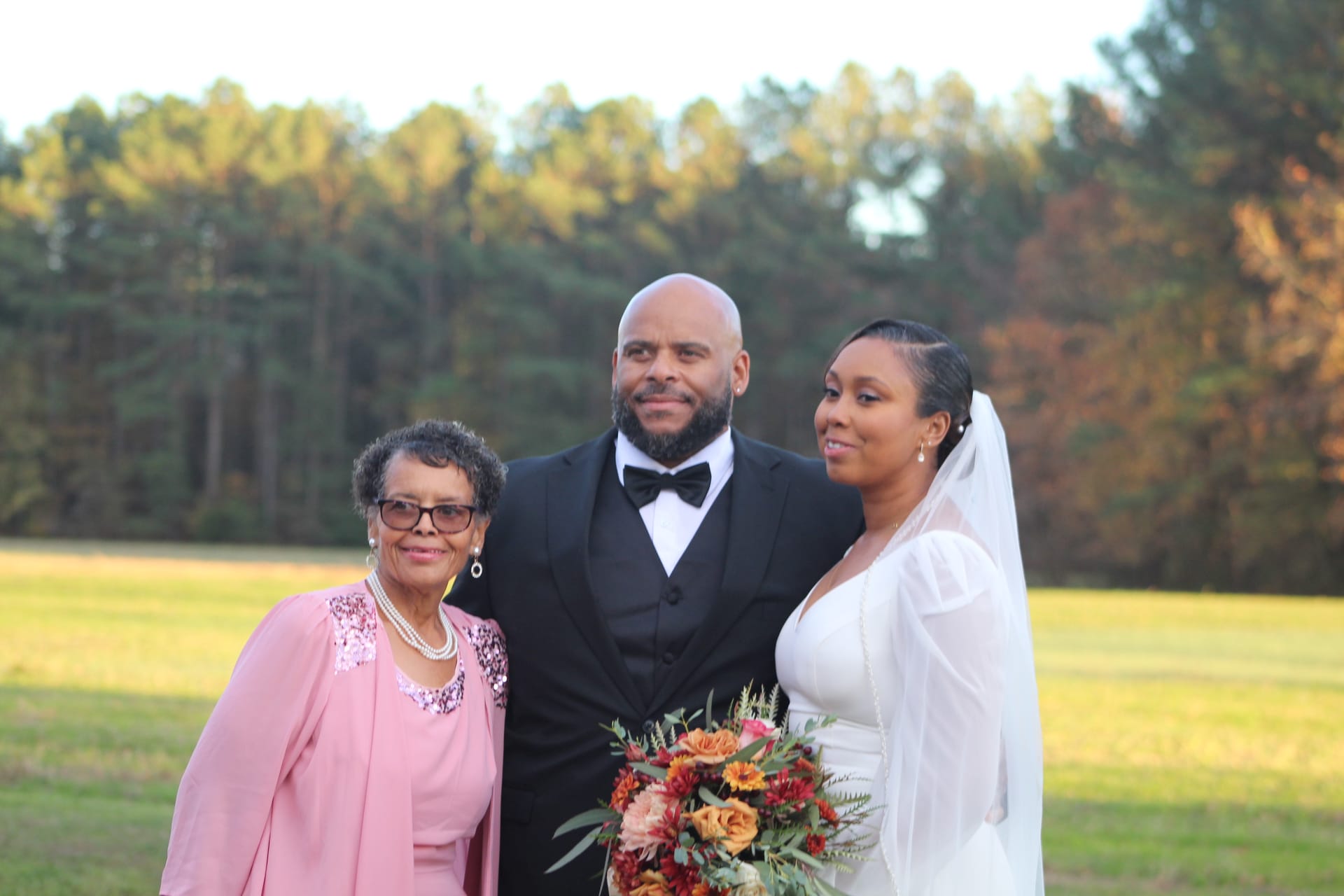 Bride in white gown holding an autumn bouquet and groom in black tuxedo pose with an elder family member on the open farmland fields at Scarborough House during golden hour.
