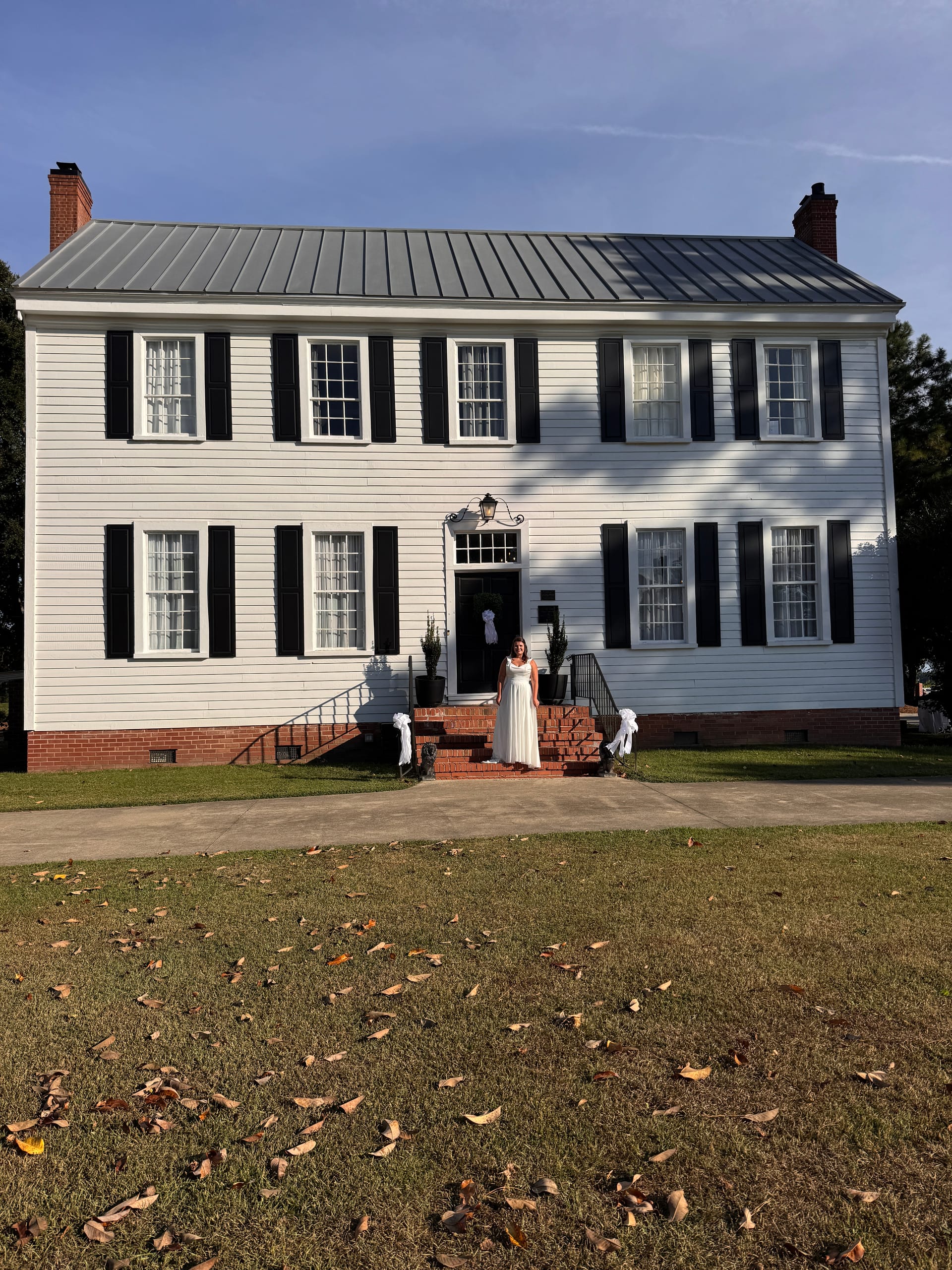 Bride in a white gown standing on the brick front steps of the 1821 Scarborough House historic manor, framed by black shuttered windows and a metal roof under a clear blue sky.
