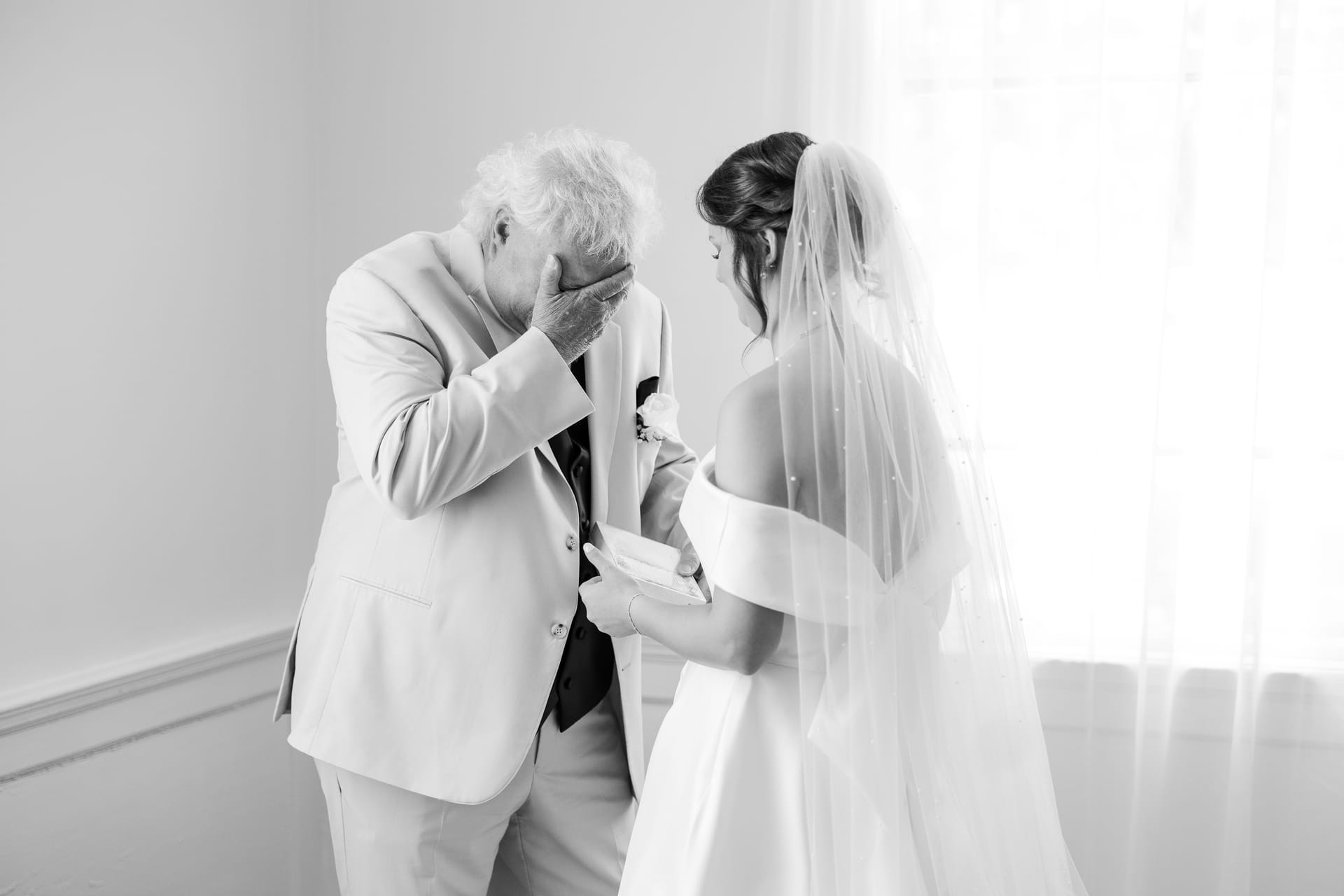 An emotional older gentleman in a suit covers his face while a bride in a white off-shoulder gown and veil holds his hands in a tender indoor moment at Scarborough House.