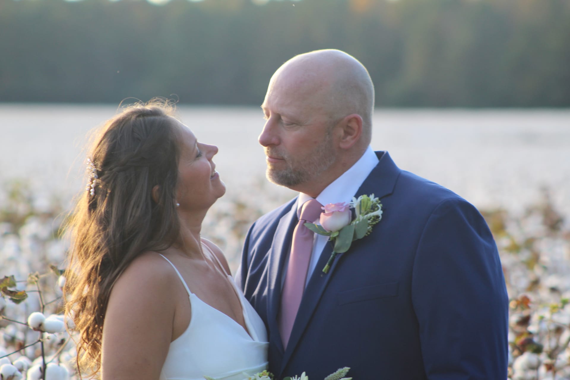 Bride in white dress and groom in navy suit with pink boutonniere share an intimate gaze in a sunlit cotton field at Scarborough House.