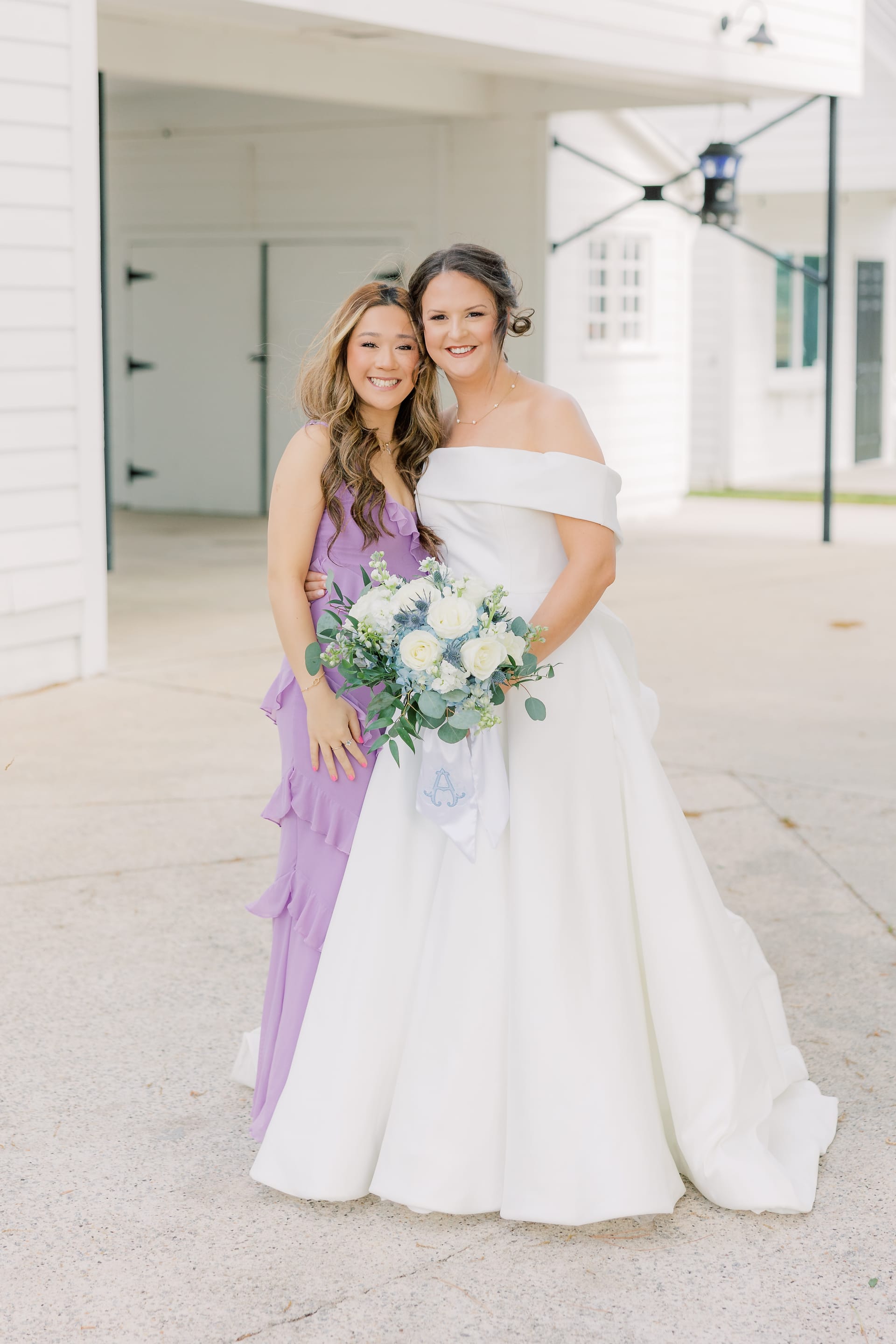 A smiling bride in an off-shoulder white gown holding a white and blue bouquet poses with a guest in a purple ruffled dress outside a white historic venue.