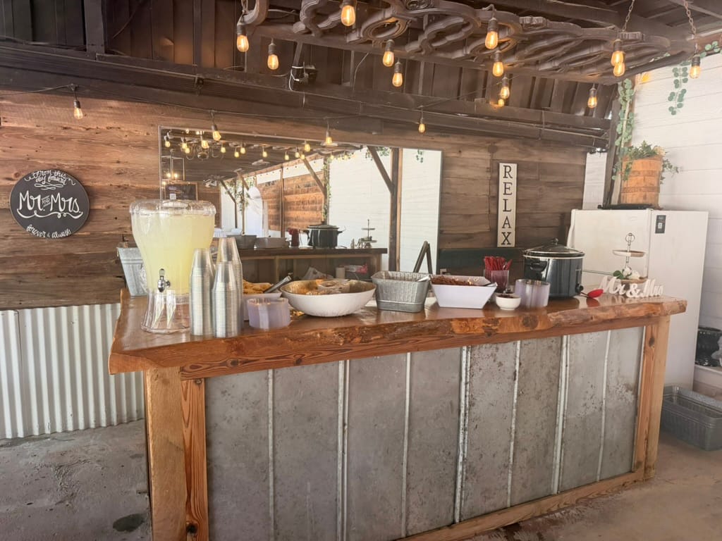Rustic live-edge wooden bar topped with a lemonade dispenser, plastic cups, food trays, and Mr and Mrs decor beneath Edison bulb string lights at a barn-style wedding venue.