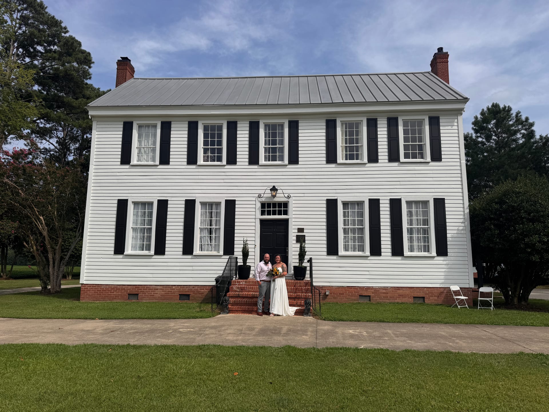 Bride in white gown and groom in gray suit pose together on the brick front steps of the 1821 Scarborough House manor under a partly cloudy blue sky.