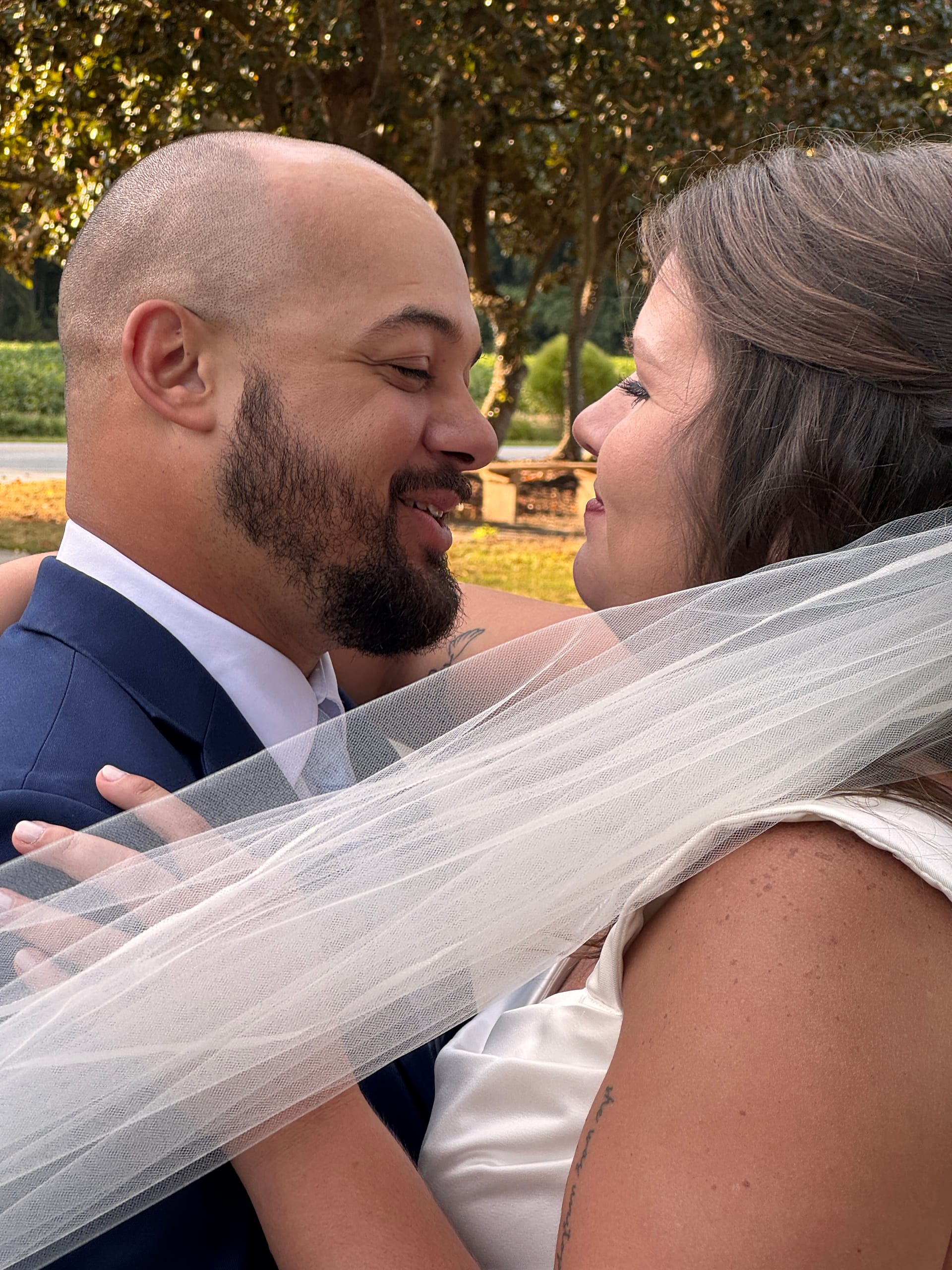 Bride and groom gazing at each other and smiling beneath oak trees at Scarborough House, the bride's veil softly draped between them in warm golden-hour light.