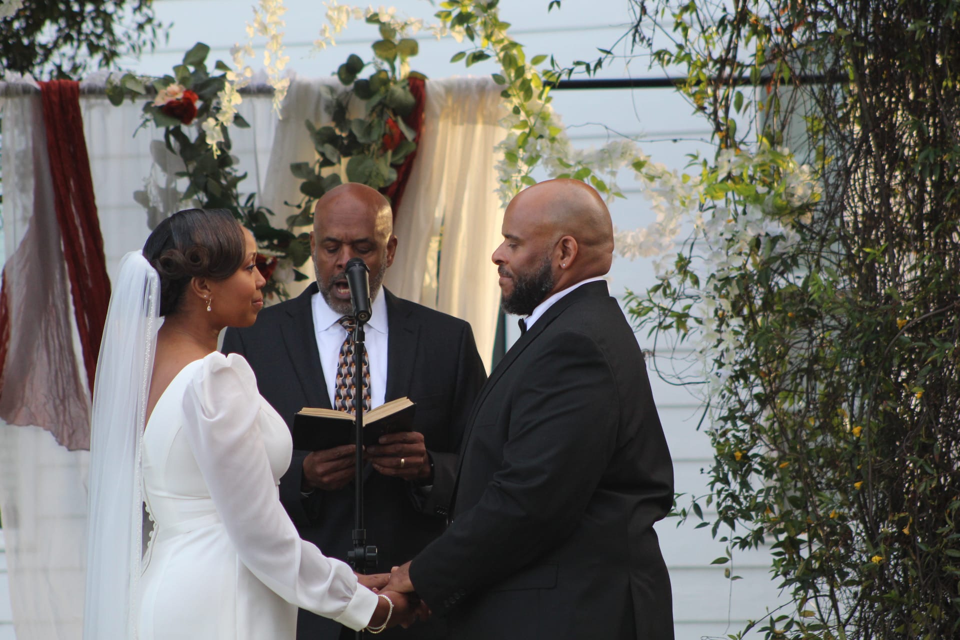 Bride in a long-sleeve white gown and groom in a black tuxedo hold hands during their outdoor wedding ceremony as the officiant reads from a book beneath a floral arch at Scarborough House.
