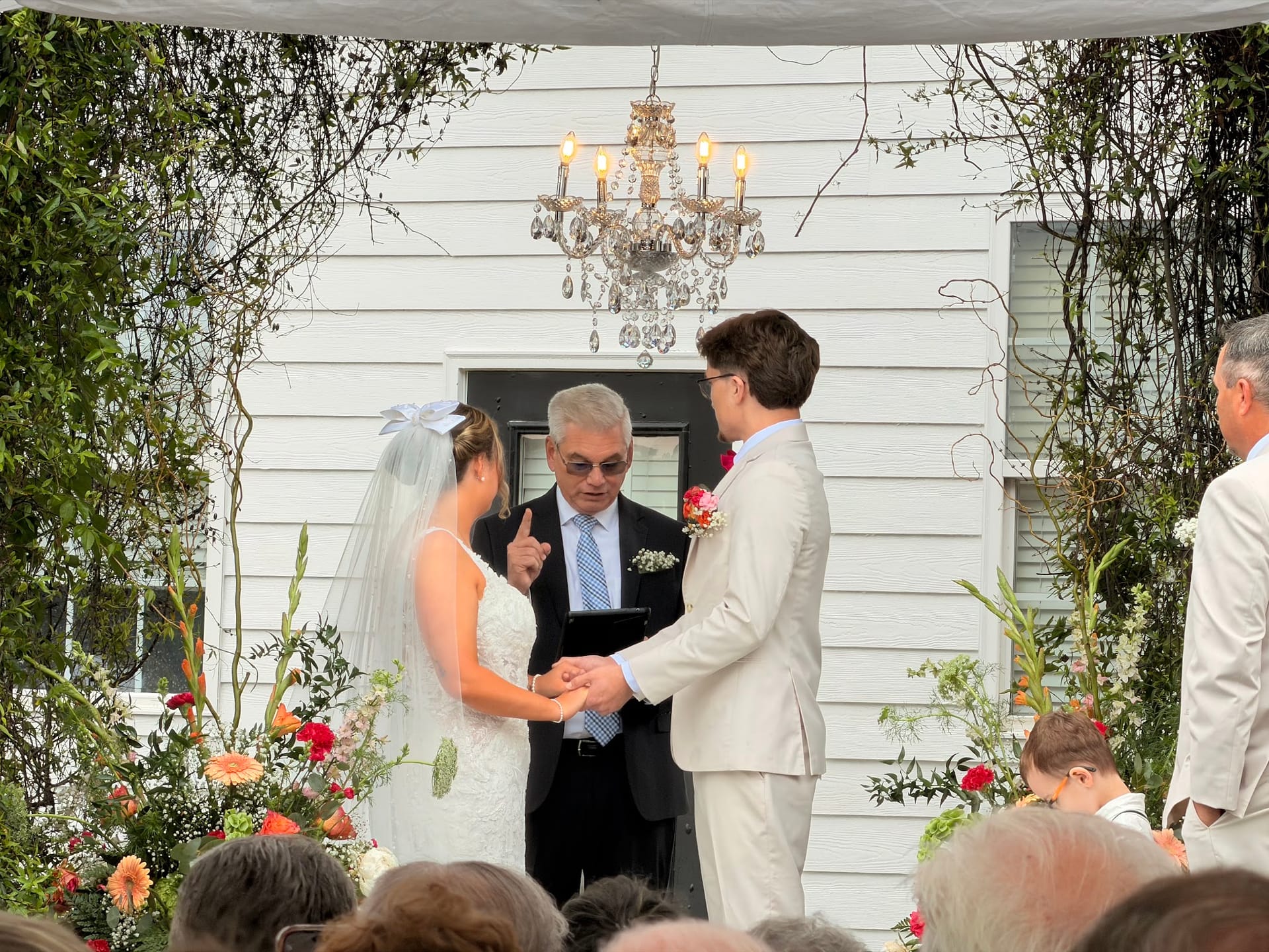 Bride and groom exchange rings during an outdoor wedding ceremony beneath a crystal chandelier at Scarborough House historic estate venue.