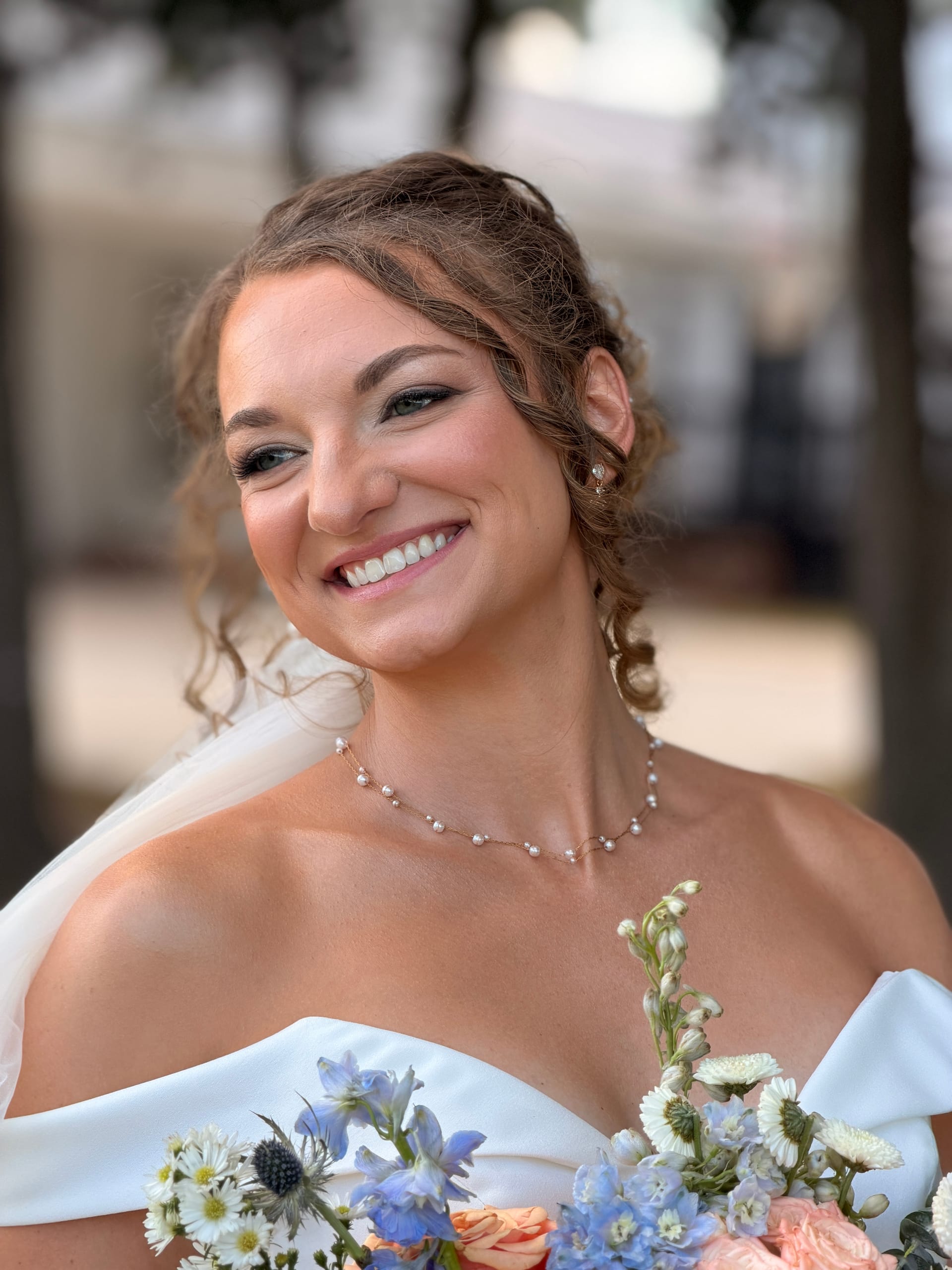 A glowing bride in an off-shoulder white gown smiles warmly while holding a pastel bouquet of blue, white, and peach flowers beneath oak trees at Scarborough House.