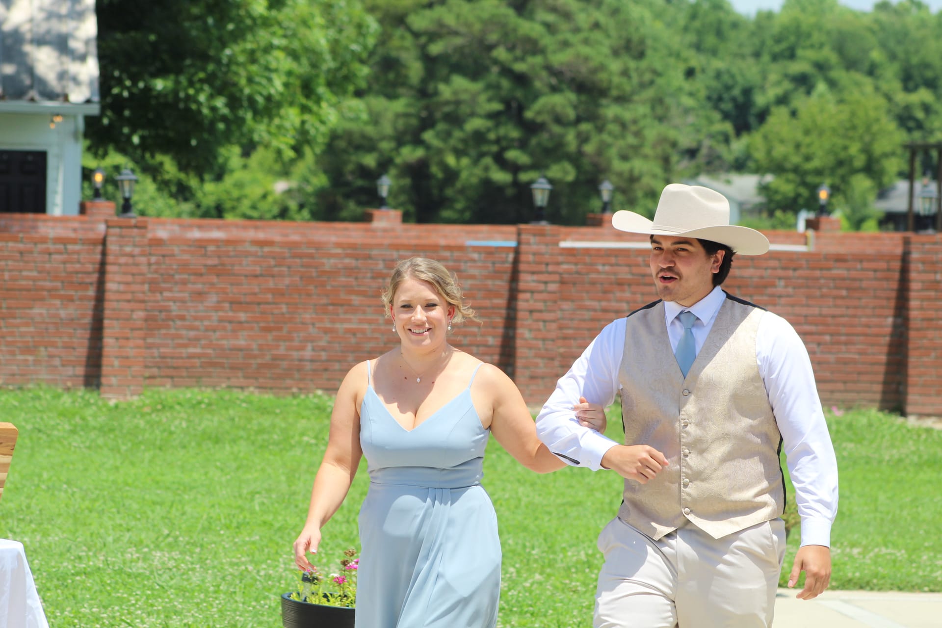 A smiling bridesmaid in a light blue gown walks arm-in-arm with a groomsman wearing a tan vest and white cowboy hat across the sunlit ceremony lawn at Scarborough House historic estate wedding venue.