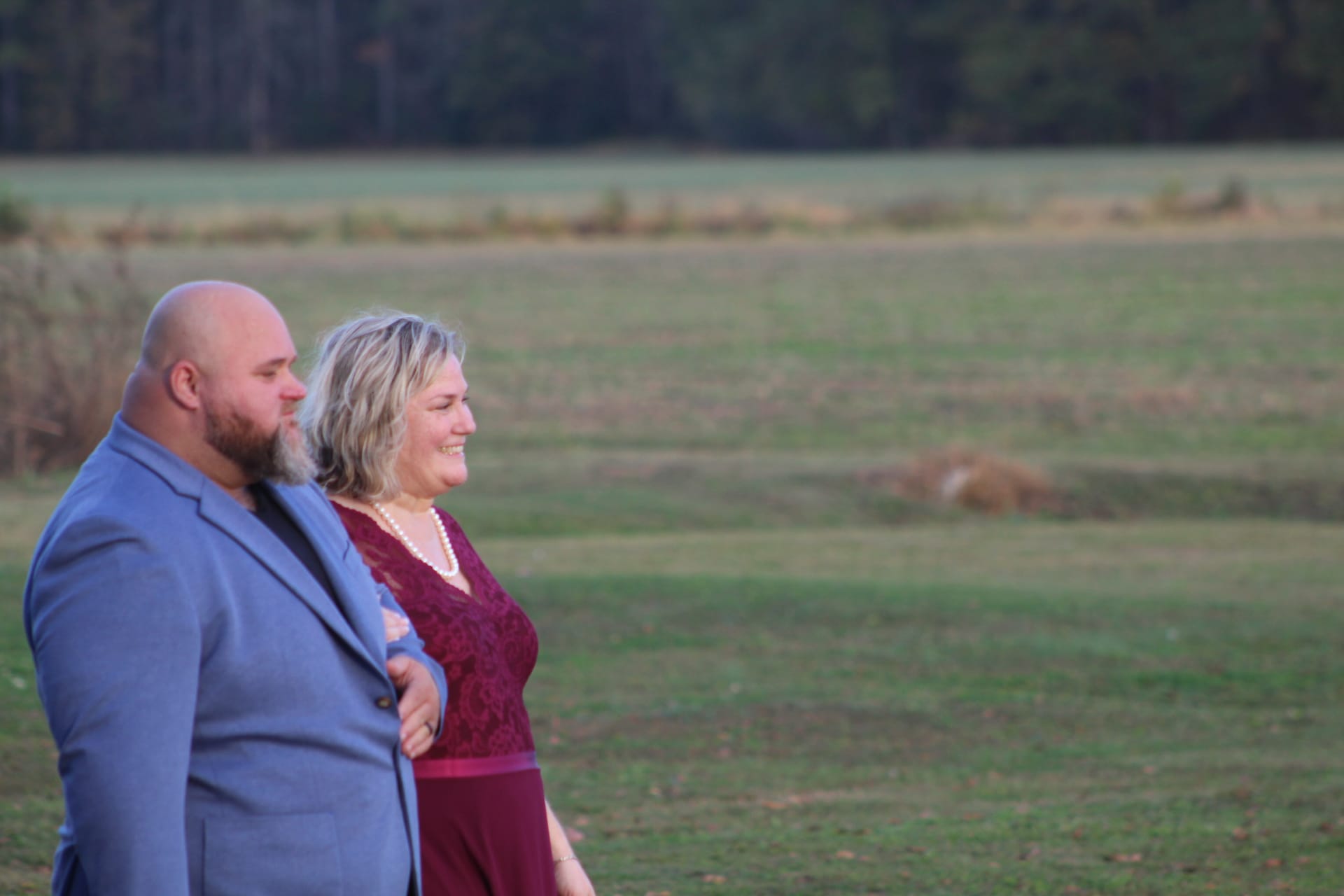 A smiling couple dressed in burgundy and blue walk arm in arm across open farmland fields at golden hour with a tree line in the background at Scarborough House estate.