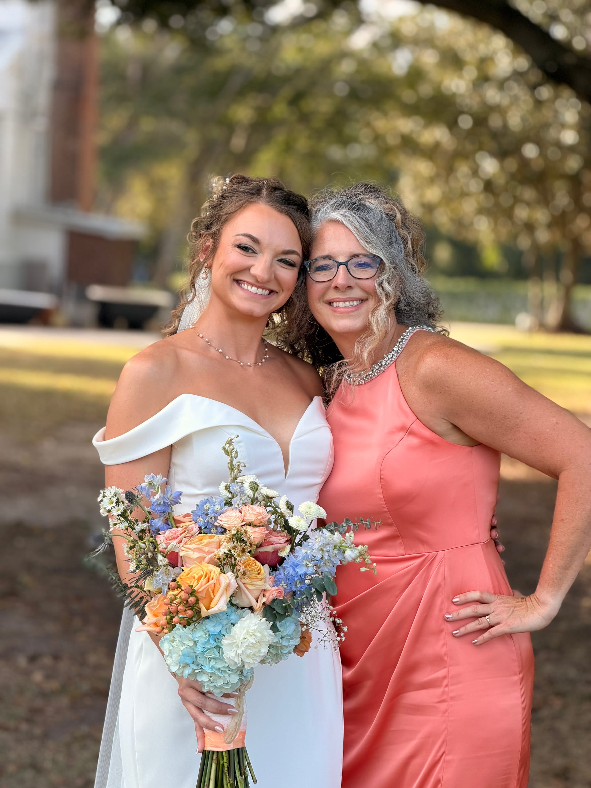 Bride in an off-shoulder white gown holding a colorful bouquet of roses and hydrangeas smiles with an older woman in a coral dress beneath the oak trees at Scarborough House historic estate.