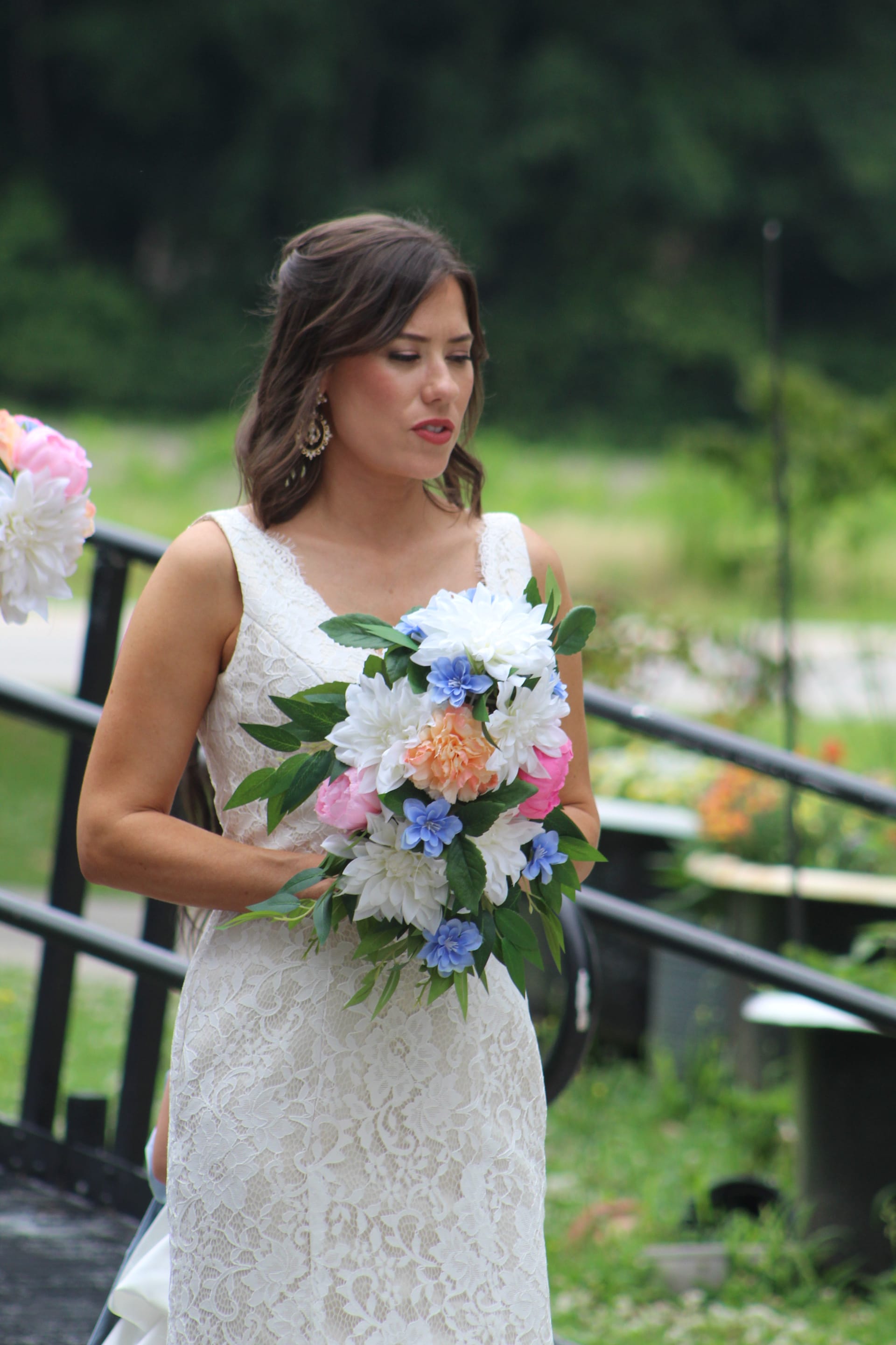 Bride in a white lace gown holding a colorful cascading bouquet of pink, white, blue, and peach flowers in an outdoor garden setting at a historic wedding venue.