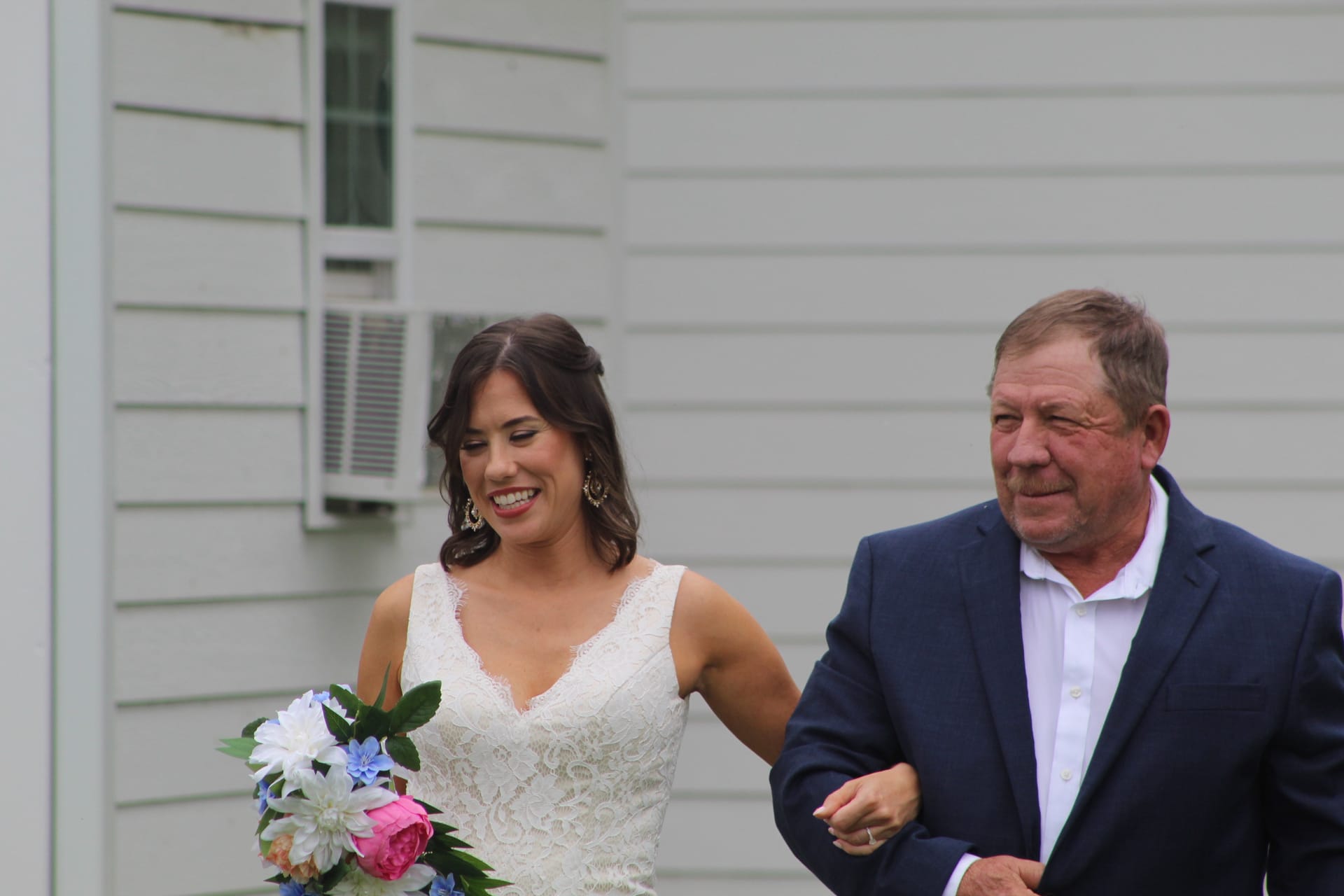 Smiling bride in a lace wedding gown holding a colorful bouquet walks arm in arm with an older gentleman in a navy blazer beside a white clapboard building.