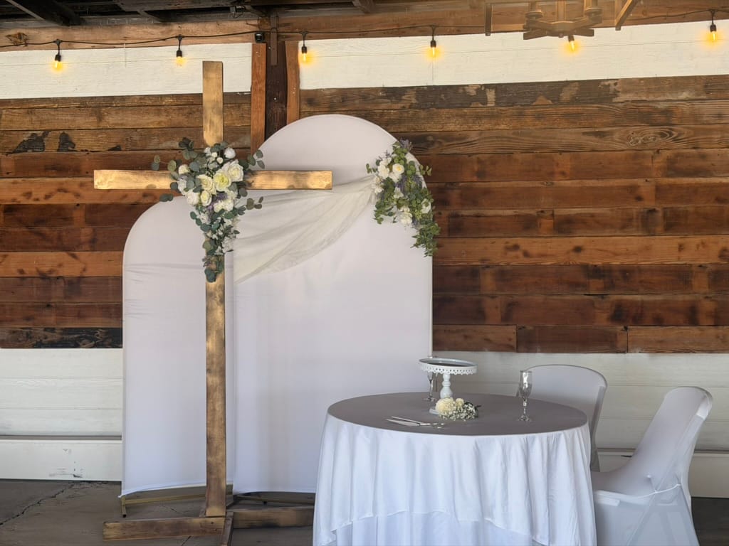 Rustic wooden cross and white arched backdrop decorated with white roses and eucalyptus greenery beside a draped signing table at Scarborough House wedding venue.