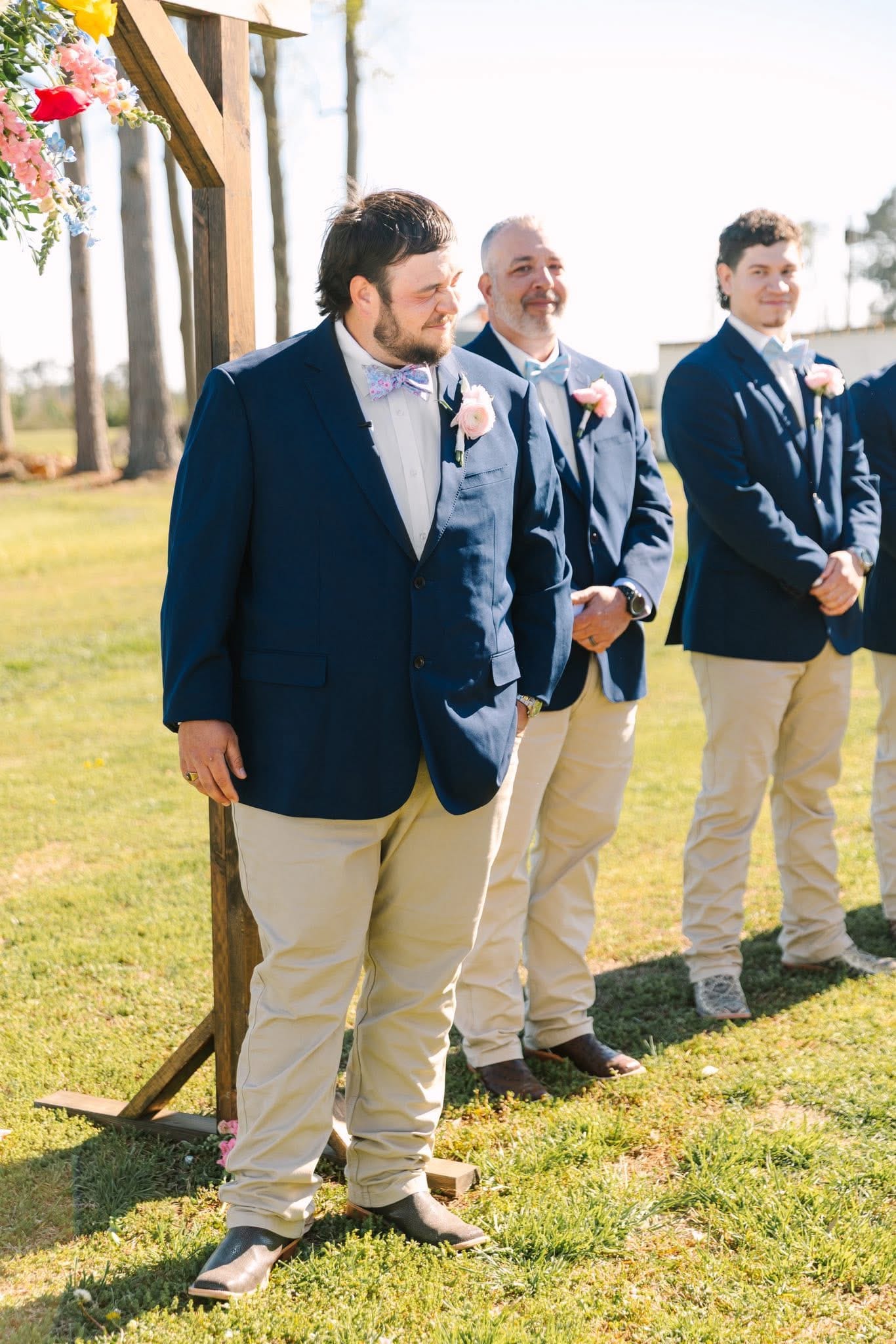 Groom in a navy blazer and floral bow tie watches the bride approach at an outdoor wedding ceremony on the Scarborough House ceremony lawn, with smiling groomsmen standing behind him beneath a rustic wooden arch adorned with colorful florals.