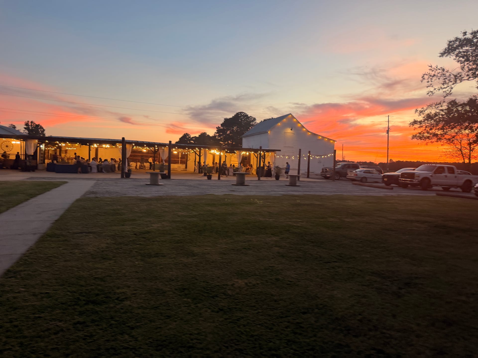 Guests gather under string lights at an outdoor pavilion beside a white barn venue during a vibrant orange and pink sunset reception at Scarborough House.