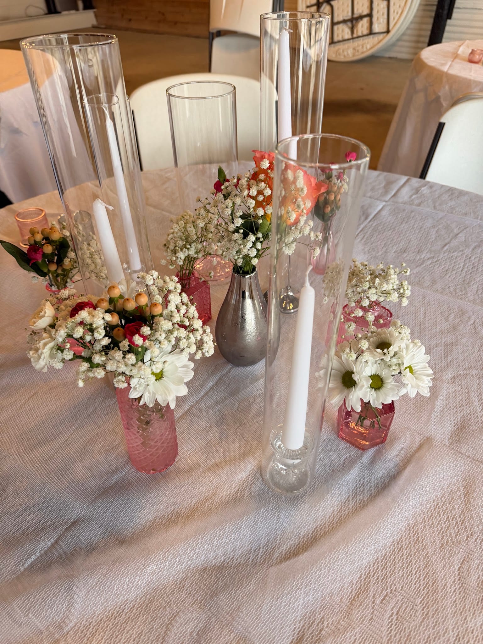 Elegant wedding reception table centerpiece featuring pink glass vases filled with baby's breath, white daisies, and coral roses alongside tall glass candle holders with white taper candles on a soft linen tablecloth.
