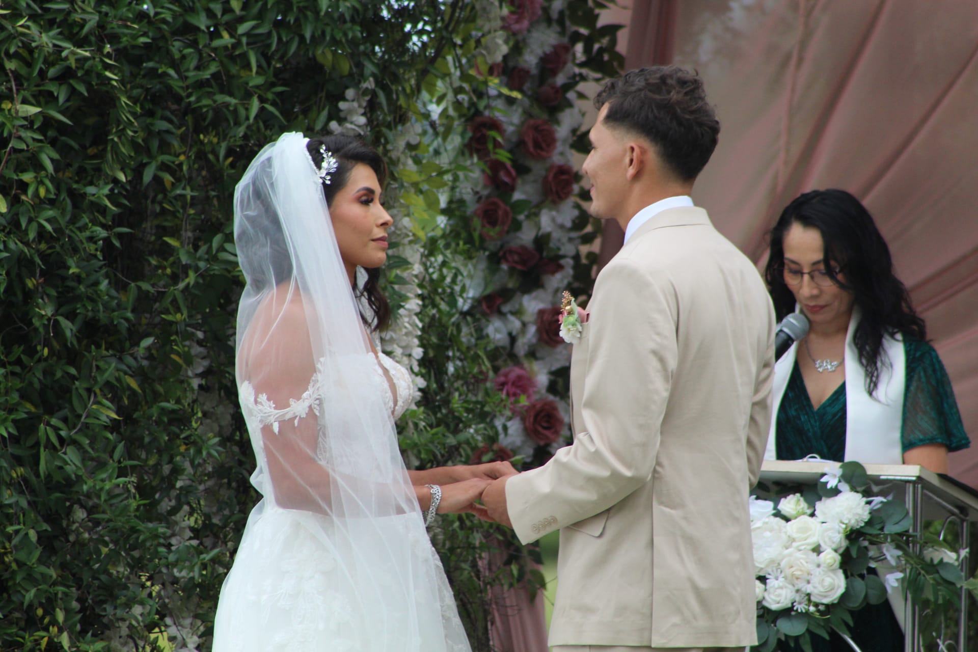 Bride in a lace ball gown and veil holds hands with groom in a tan suit during their outdoor wedding ceremony in front of a lush floral arch.