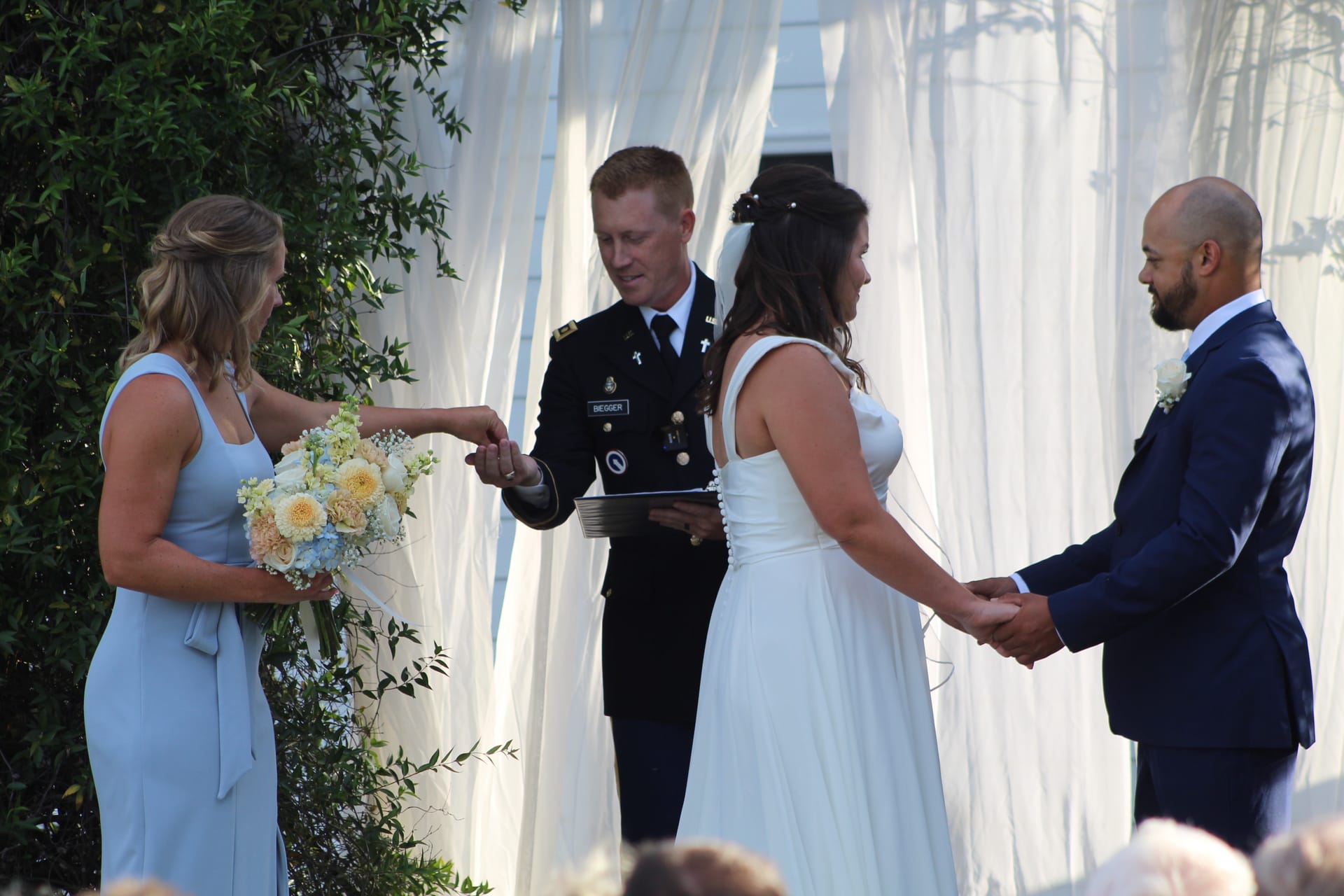 Bride and groom hold hands during their outdoor wedding ceremony officiated by a military chaplain as the maid of honor passes the ring beneath a draped white altar.