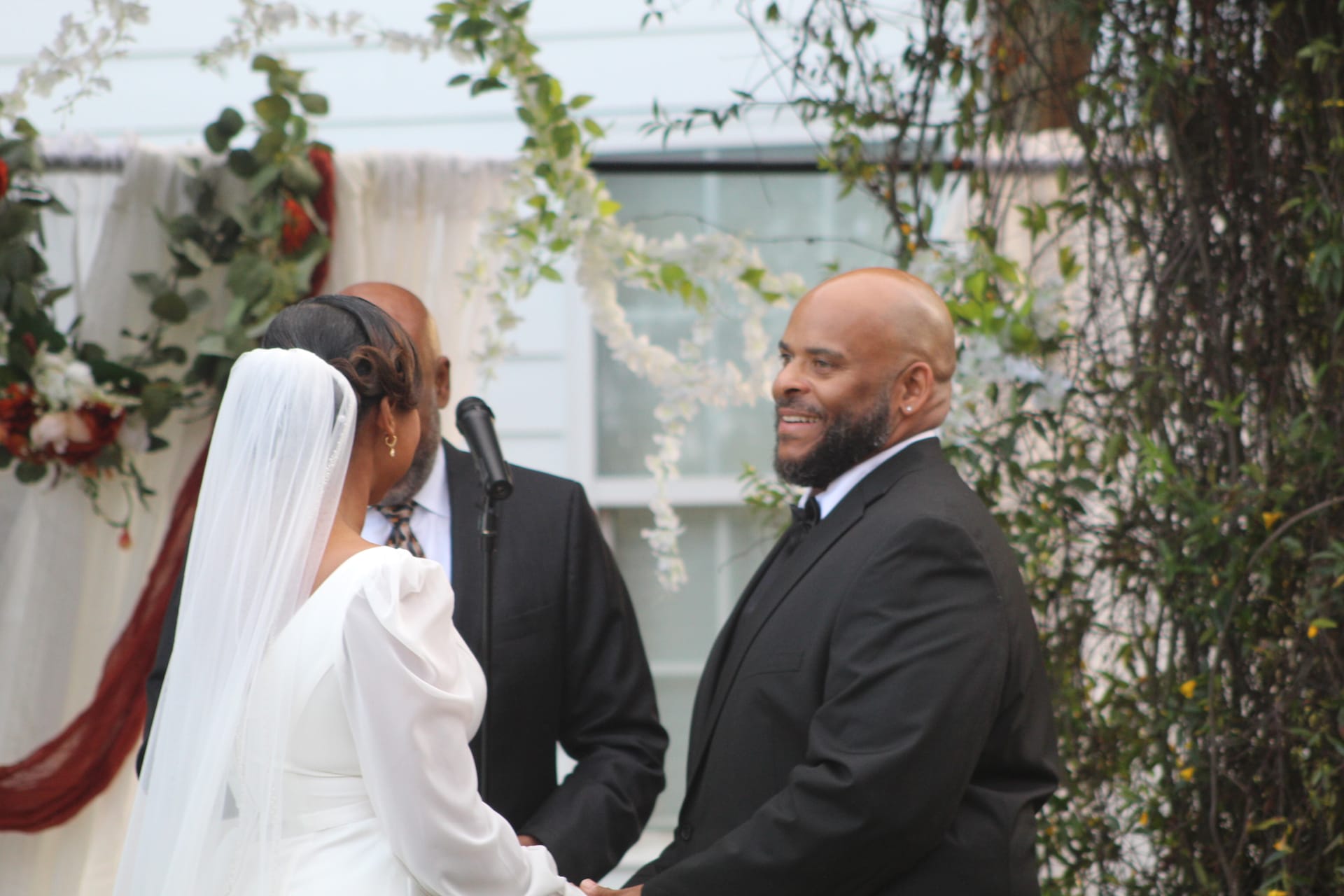 Smiling groom in a black suit holds hands with his bride in a white gown and veil during an outdoor wedding ceremony beneath a floral arch at Scarborough House.