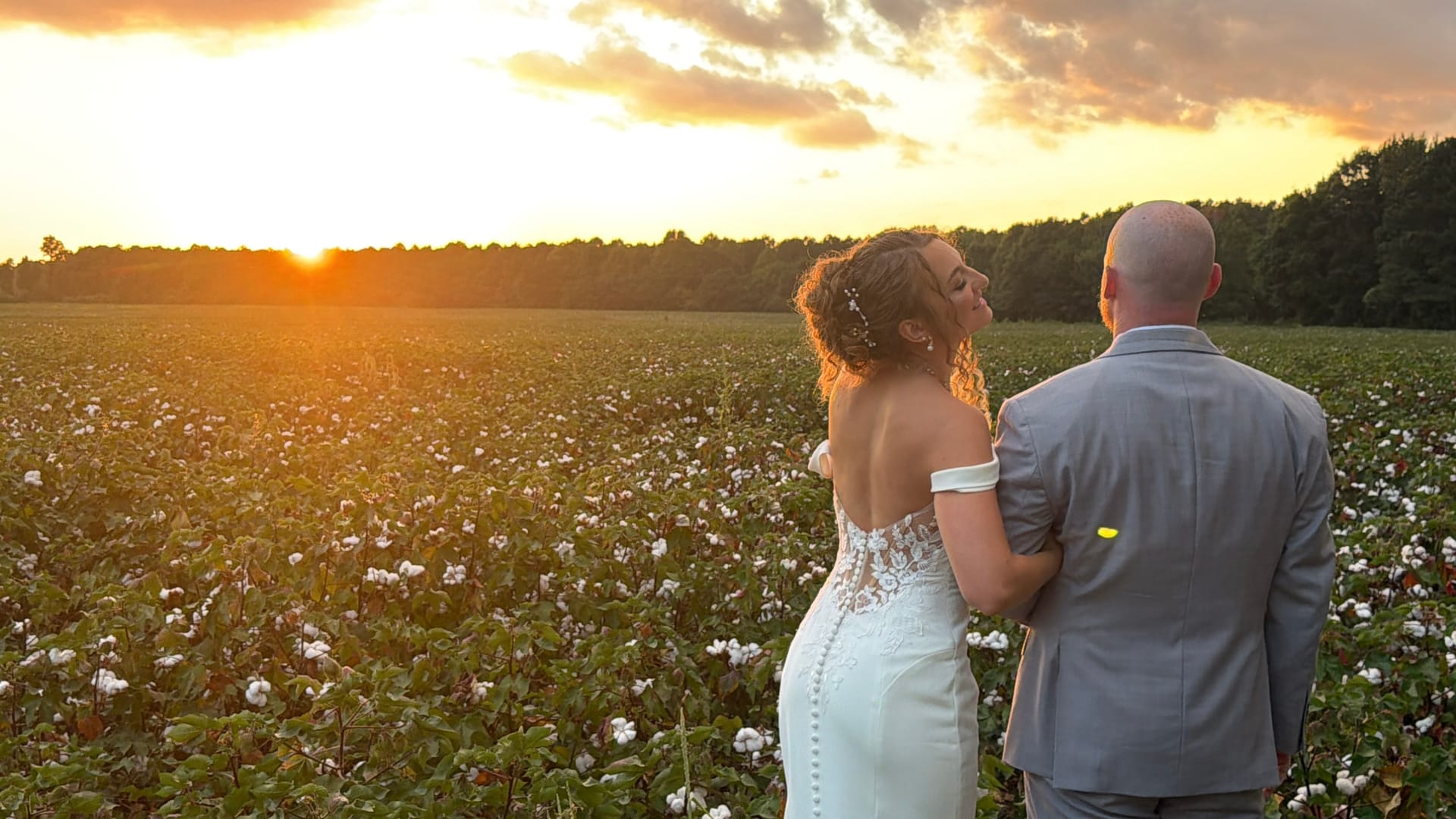 Bride in a lace off-shoulder gown and groom in a gray suit embrace while gazing toward a glowing sunset over a blooming cotton field at Scarborough House.
