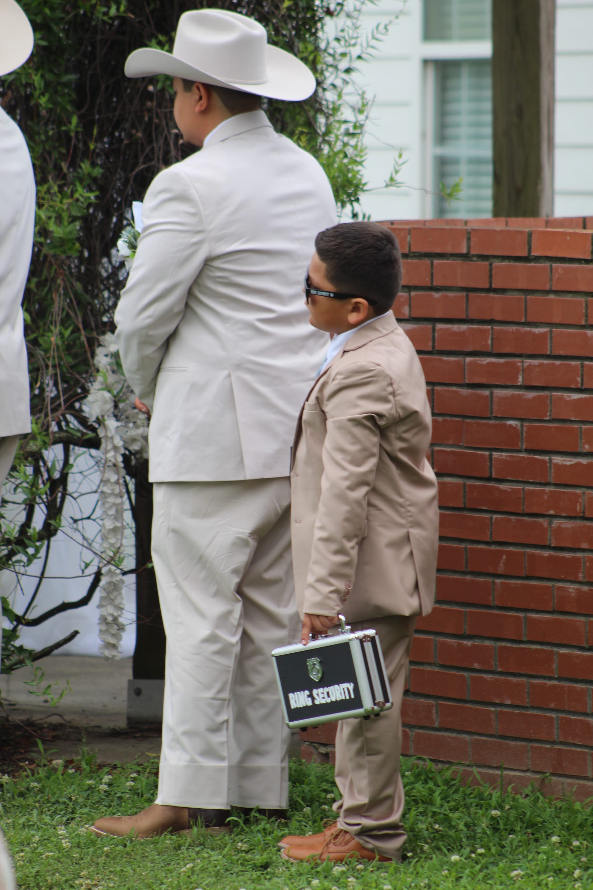 A young ring bearer in a tan suit and sunglasses holds a Ring Security briefcase while standing beside a groomsman in a white suit and cowboy hat during an outdoor wedding ceremony.