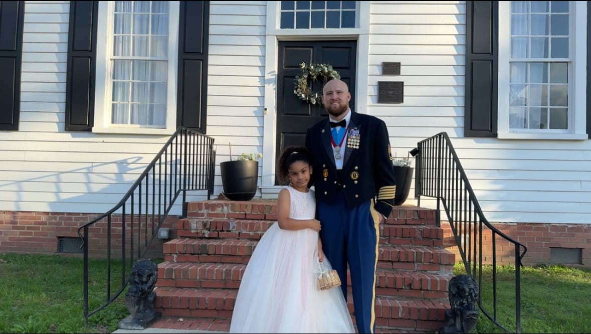 A uniformed military officer adorned with medals poses with a young girl in a white tulle gown on the brick front steps of the 1821 Scarborough House manor.