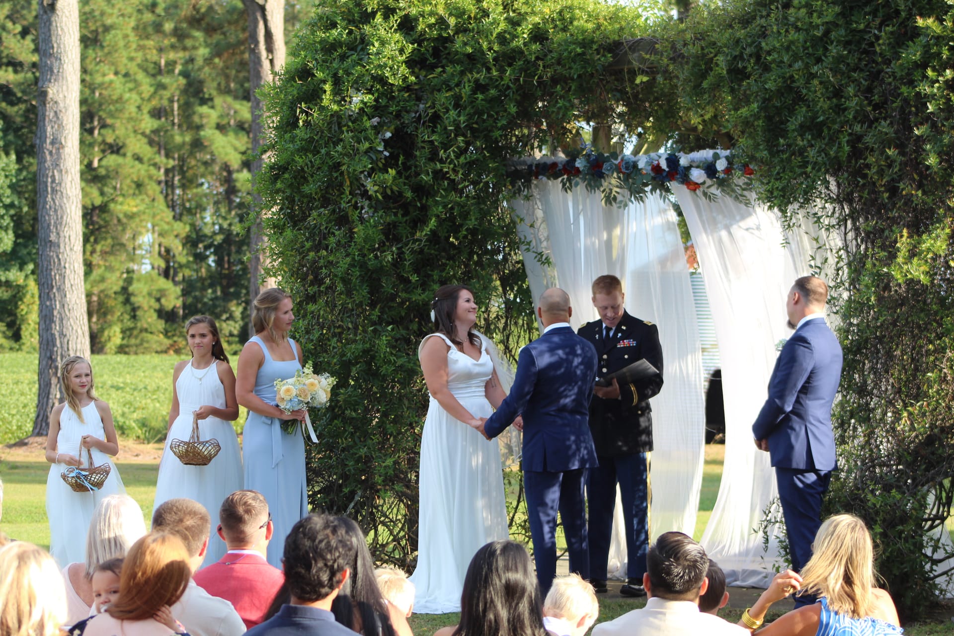 Bride and groom exchange vows beneath a lush greenery arch adorned with white draping and floral accents on the ceremony lawn at Scarborough House, with bridesmaids in light blue and a military officiant presiding.