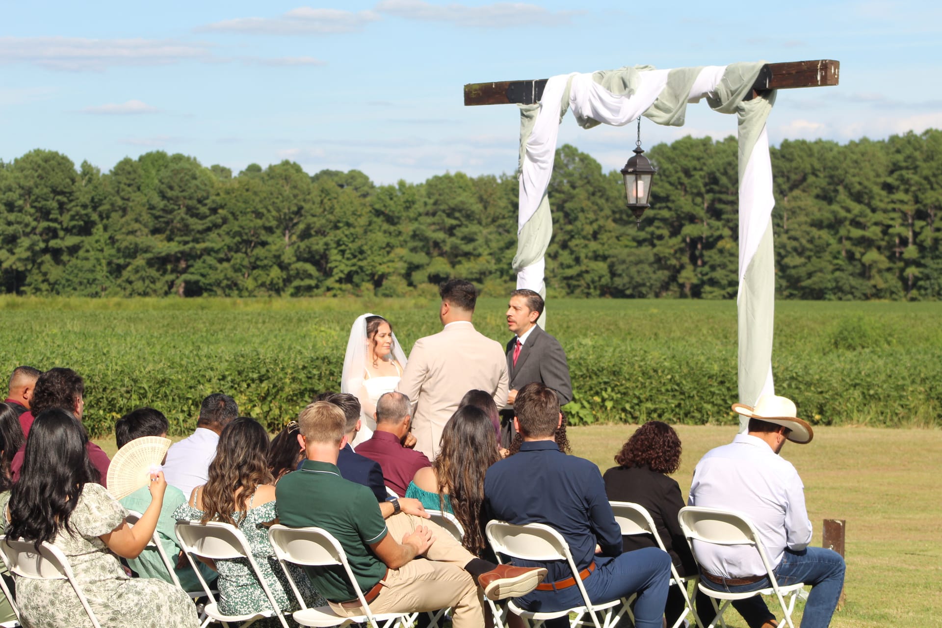 Bride and groom exchange vows beneath a draped wooden arch on the ceremony lawn at Scarborough House, surrounded by seated guests with sweeping green farmland fields and trees in the background.