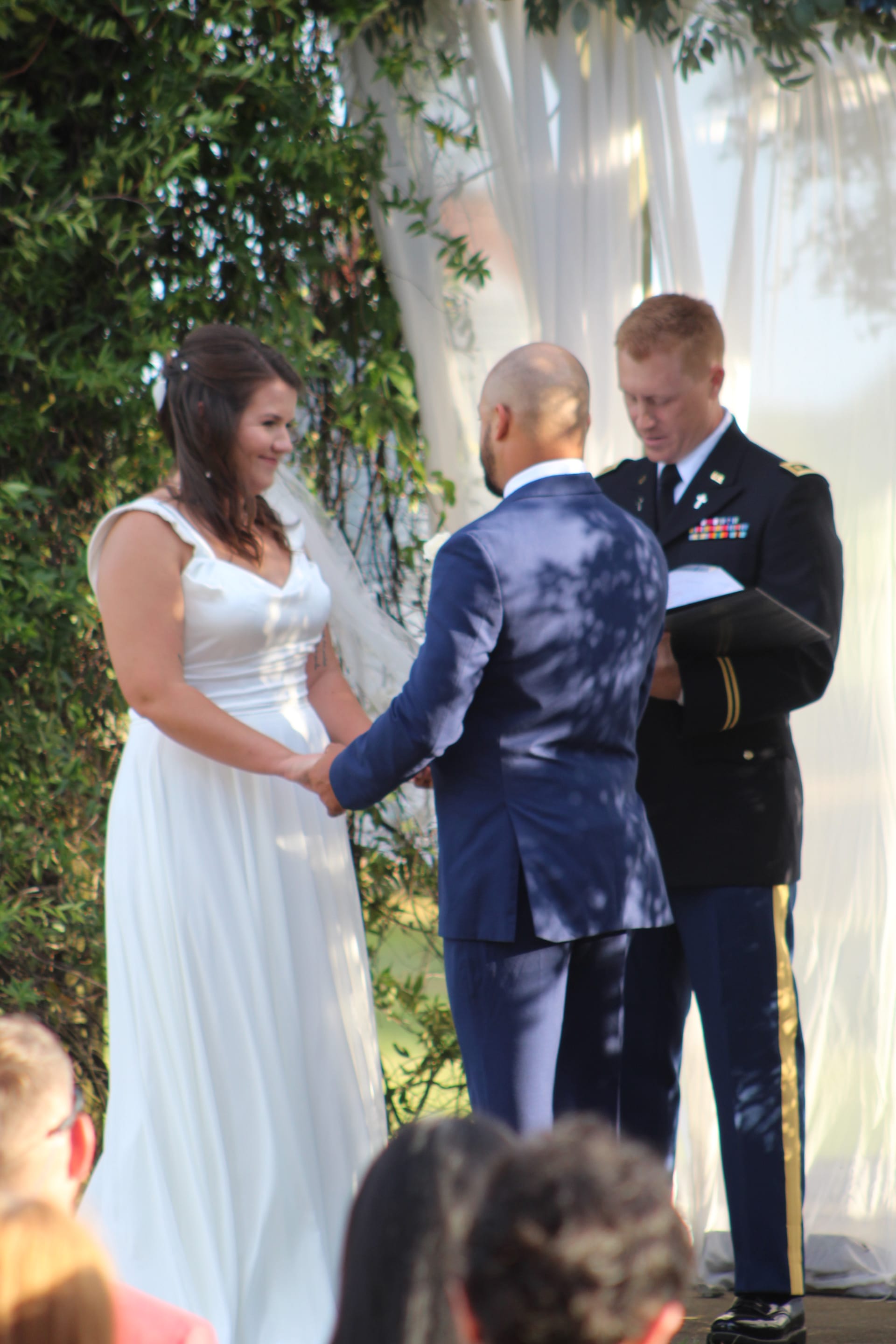 Bride in a flowing white gown smiles as she holds hands with her groom during an outdoor wedding ceremony officiated by a military officer at Scarborough House estate.