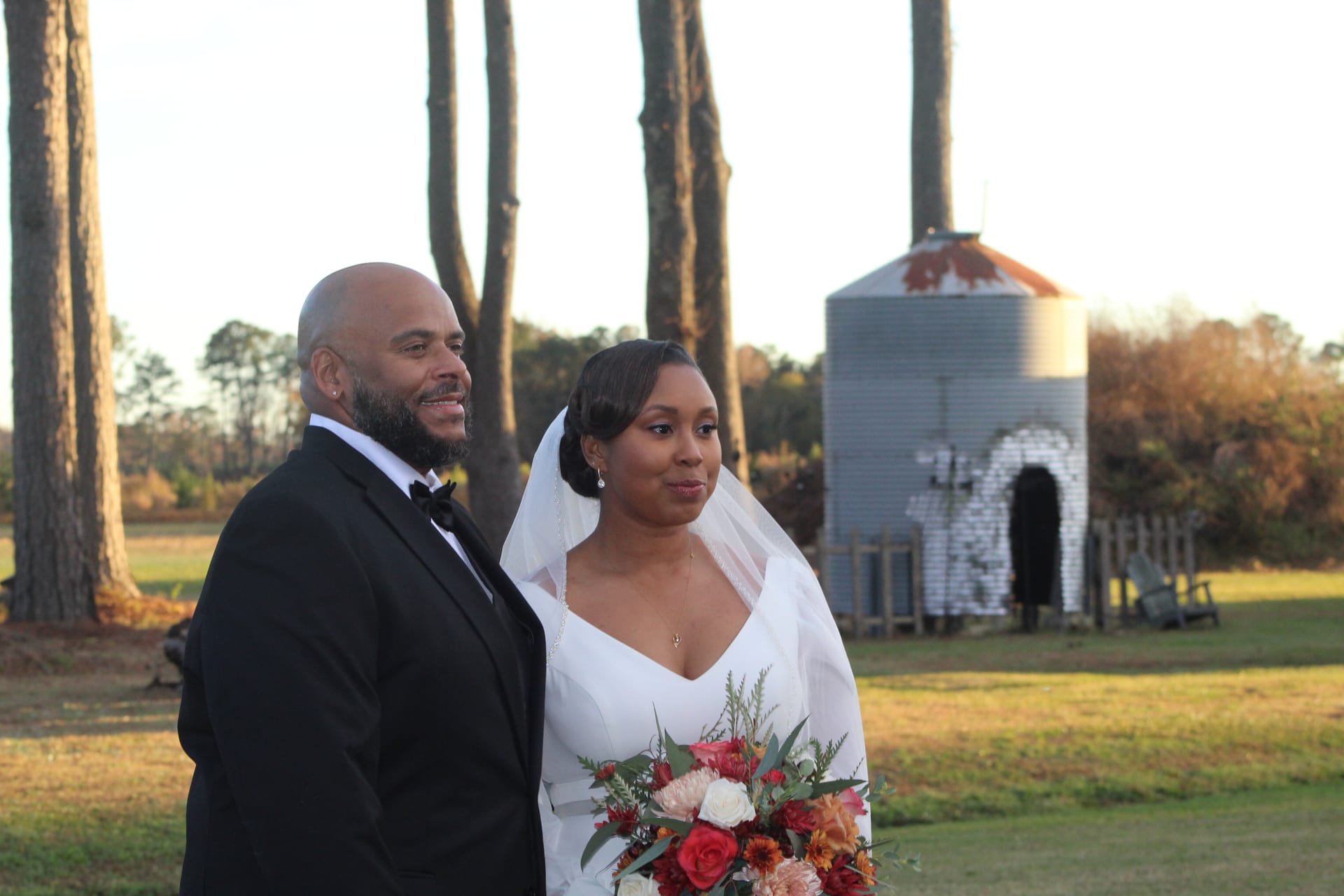 Bride holding a vibrant autumn bouquet of red and orange blooms stands beside a smiling groom in a black tuxedo during golden-hour portraits on the open farmland fields at Scarborough House historic estate wedding venue.