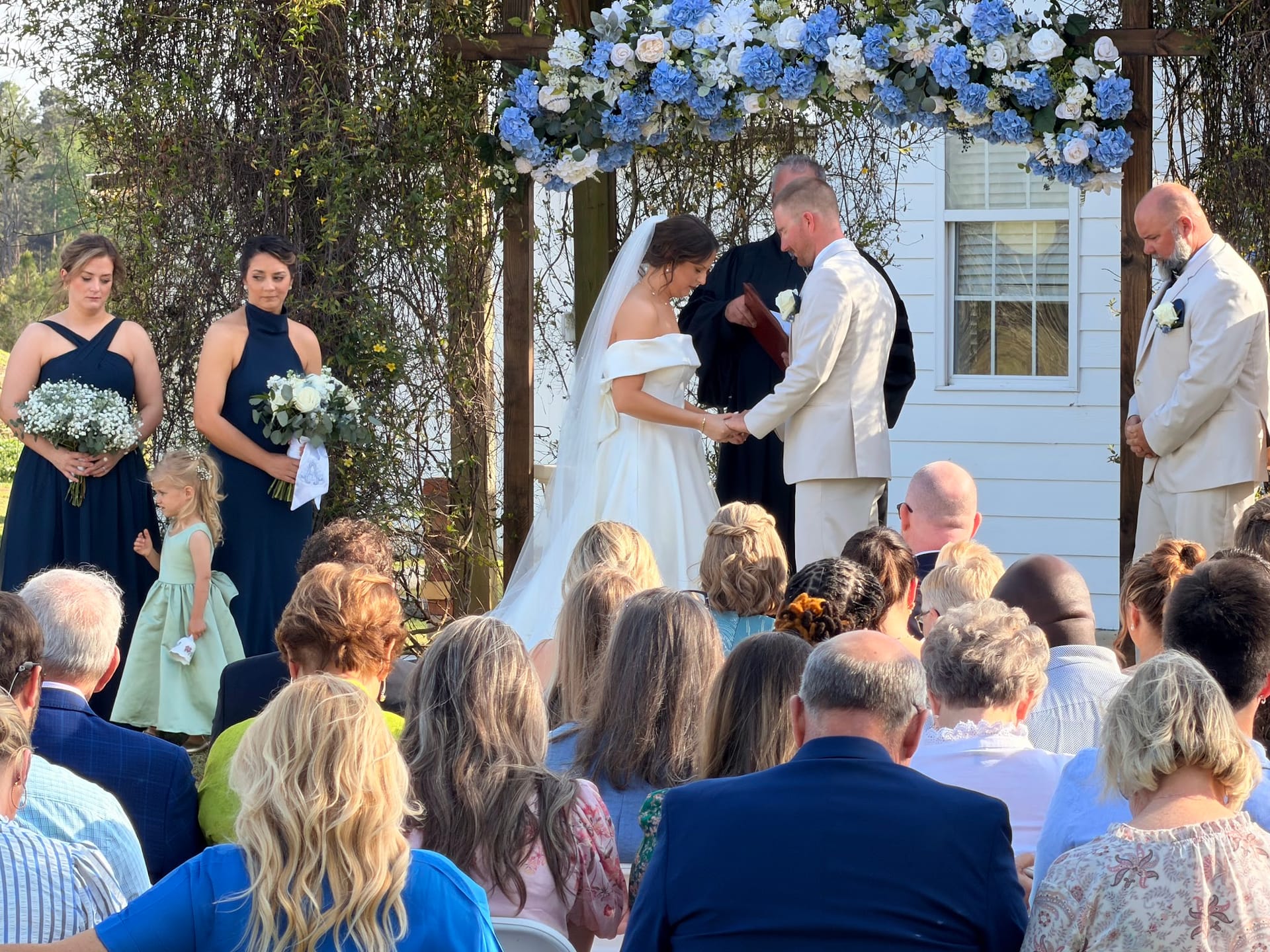 Bride and groom exchange vows beneath a lush blue and white hydrangea floral arch at an outdoor garden ceremony while guests and the wedding party look on.