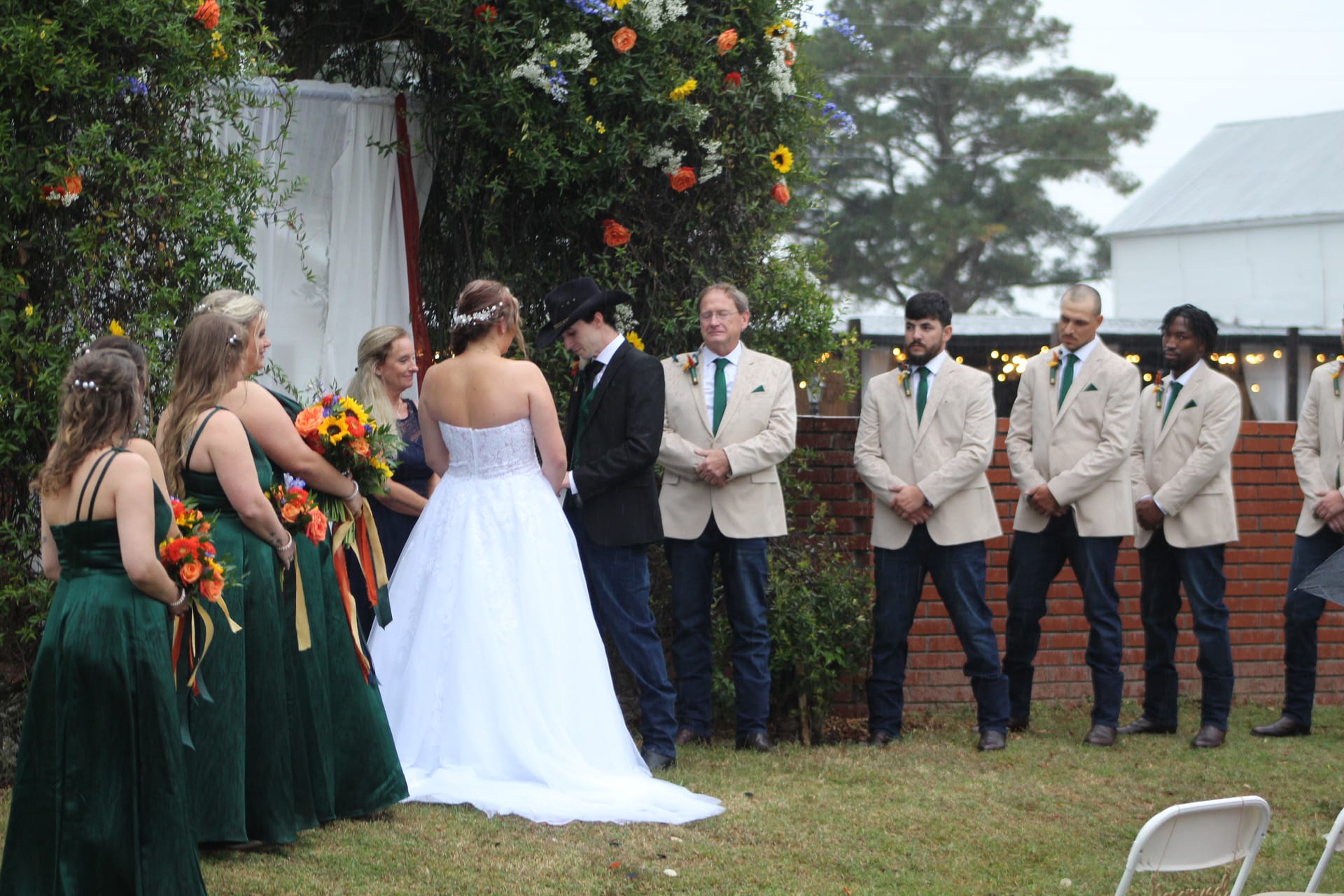 Bride in white strapless gown and groom in black hat exchange vows beneath a floral arch surrounded by bridesmaids in emerald green and groomsmen in tan blazers on the ceremony lawn at Scarborough House.