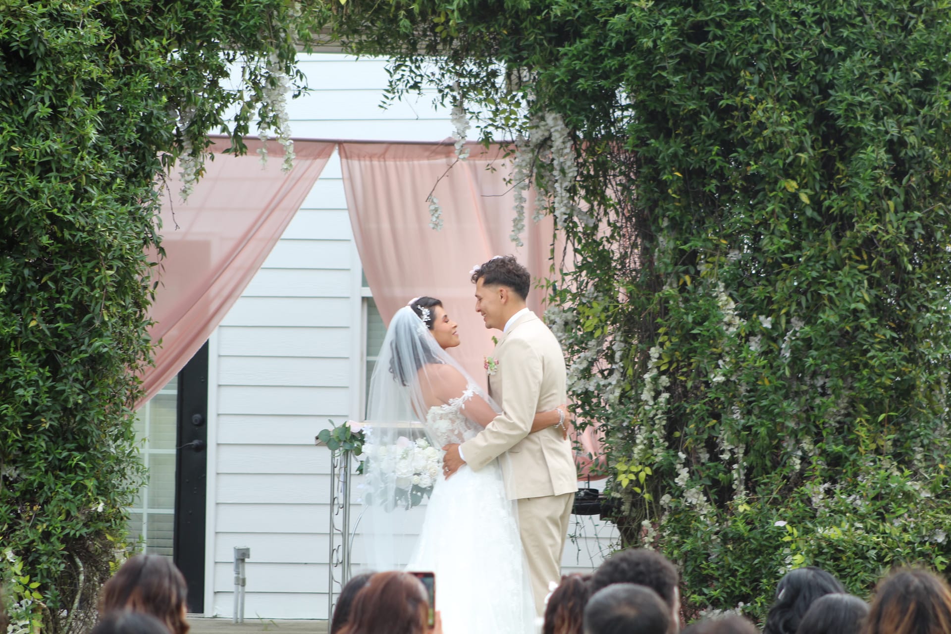 Bride in white gown and veil and groom in tan suit exchange vows at an outdoor garden ceremony framed by lush green trees and blush pink draping at Scarborough House.