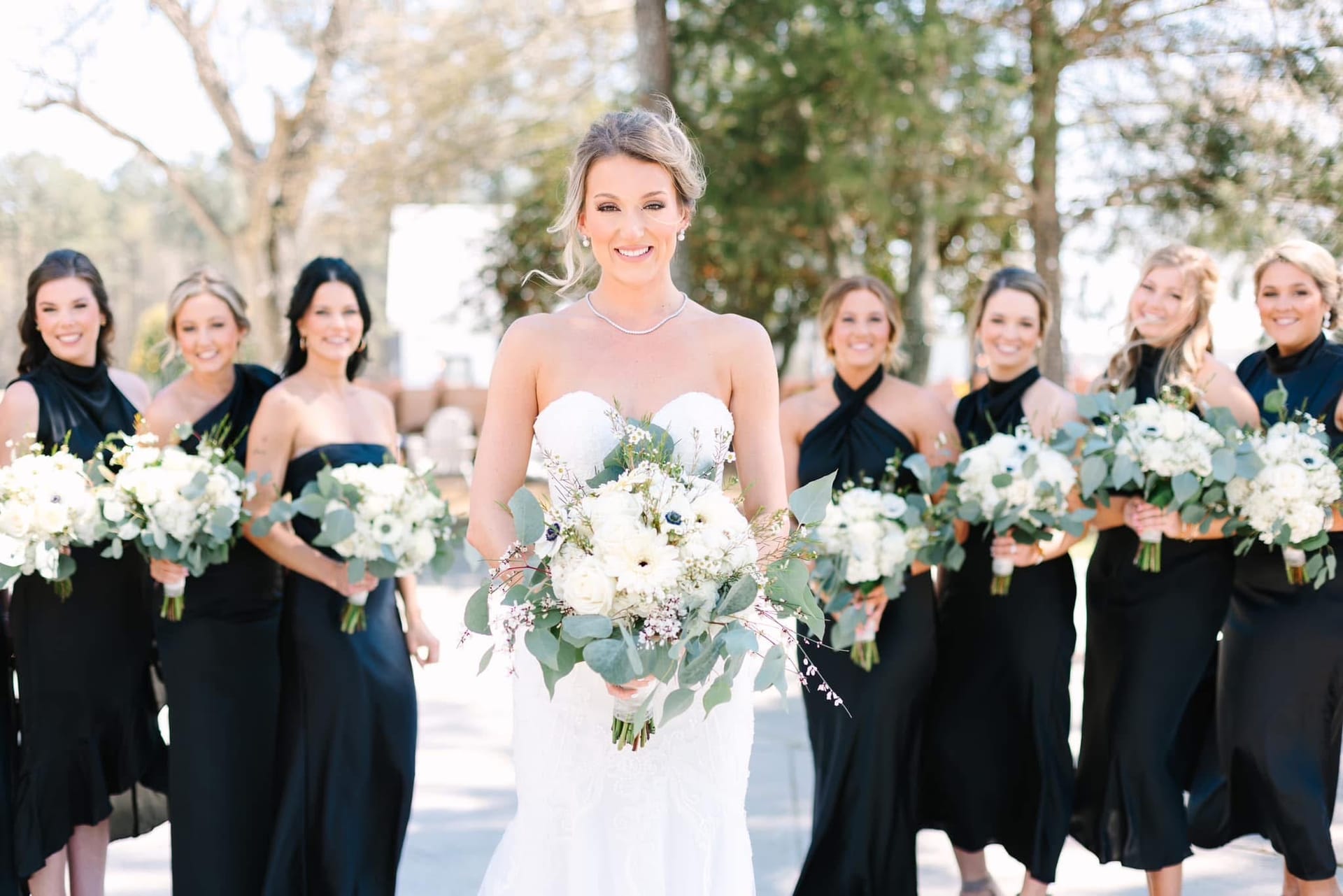 Smiling bride in a strapless white gown holds a lush white and greenery bouquet while her bridesmaids in black gowns walk behind her on the grounds of Scarborough House historic estate.