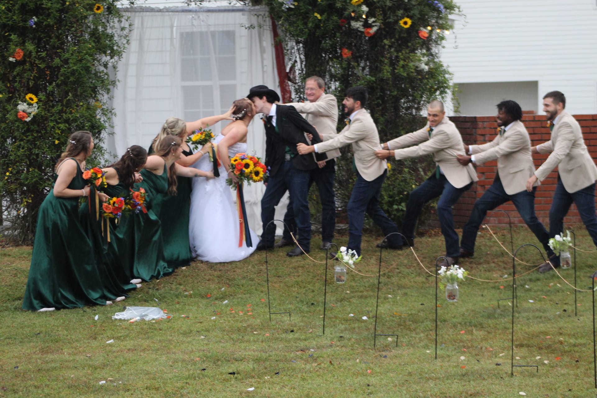 Bridesmaids in emerald green gowns and groomsmen in tan blazers playfully pull the bride and groom apart during their first kiss on the ceremony lawn at Scarborough House.