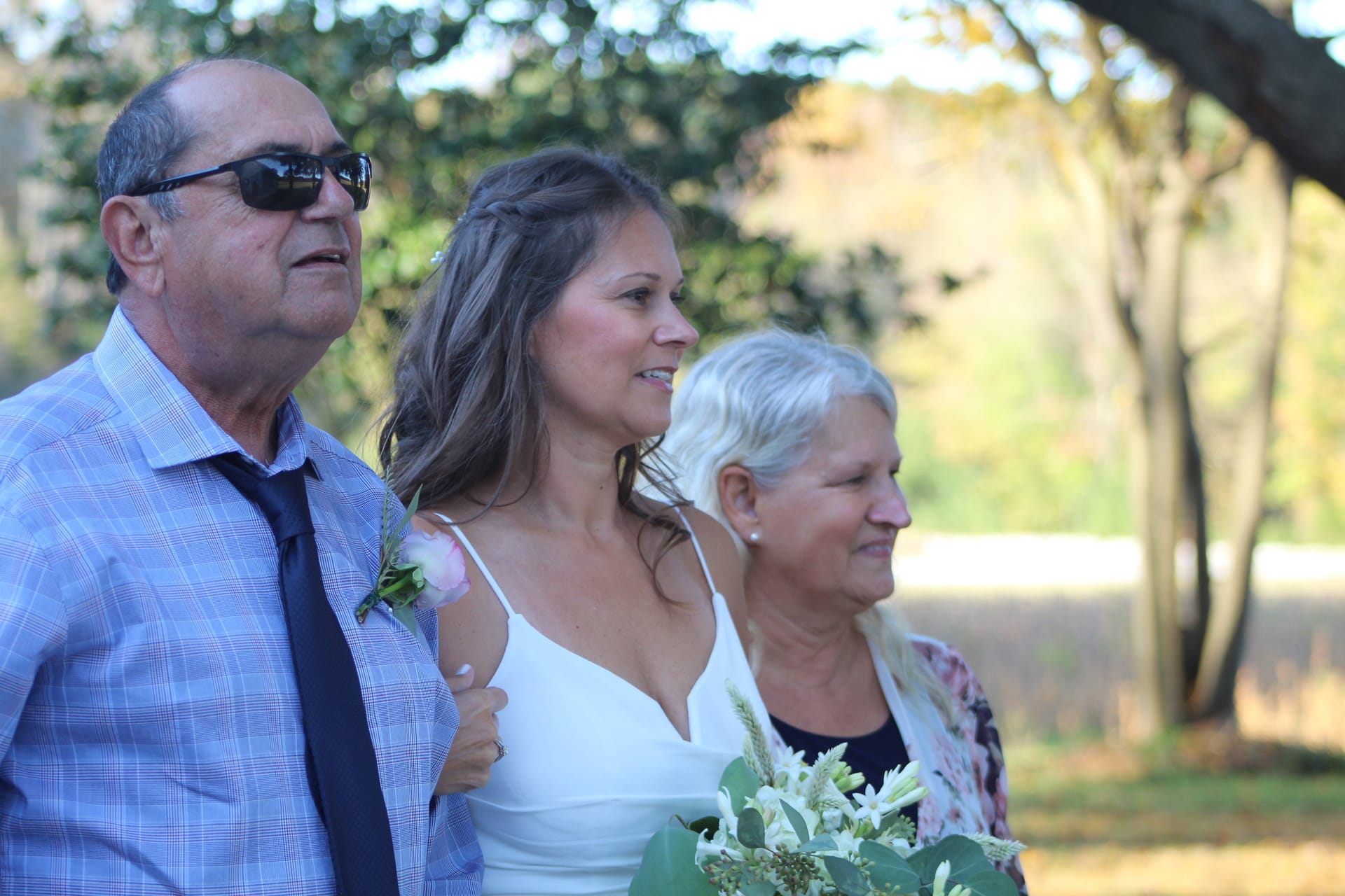 Bride in a white gown walks down the aisle flanked by her father in sunglasses and tie and her silver-haired mother holding a white floral bouquet beneath oak trees at Scarborough House.