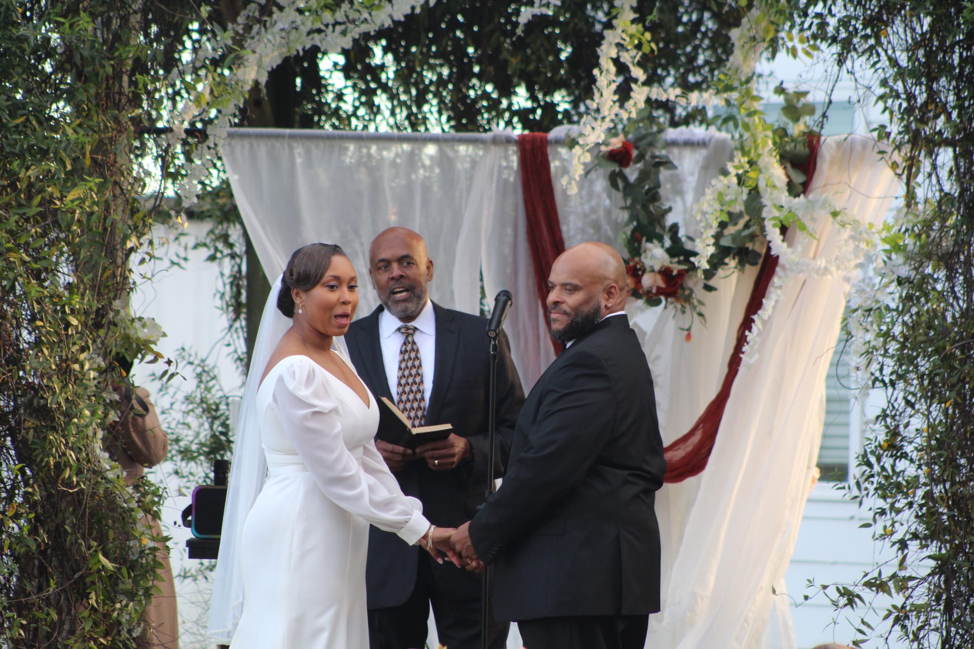Bride reciting vows to her smiling groom while holding hands beneath a draped floral arch adorned with white flowers and burgundy fabric at an outdoor wedding ceremony.