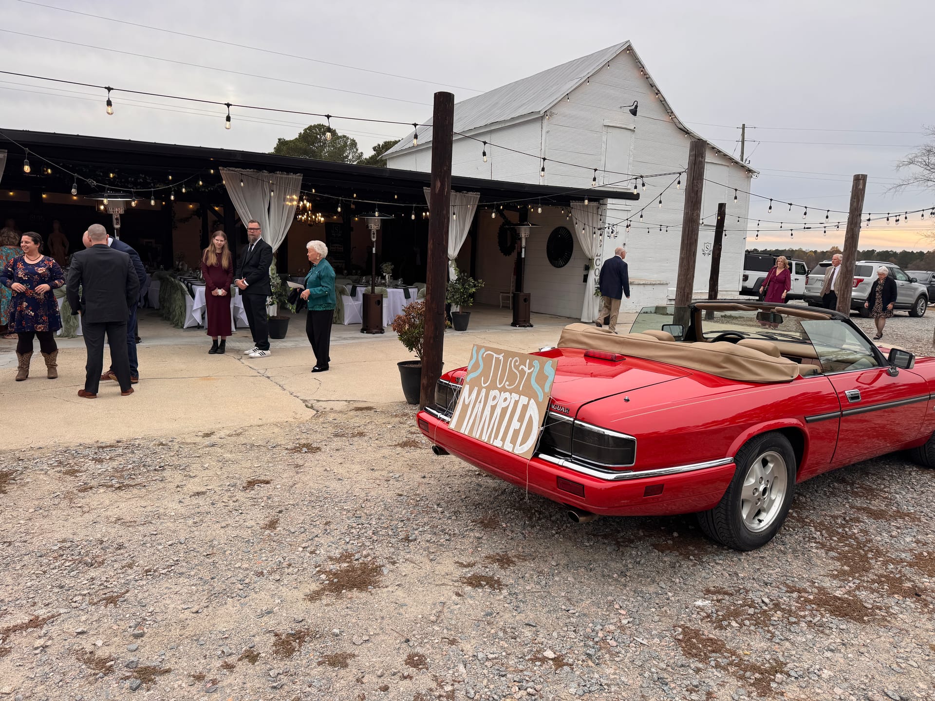 A red convertible with a Just Married sign parked in front of a white barn wedding venue decorated with string lights at dusk as guests mingle in the background.