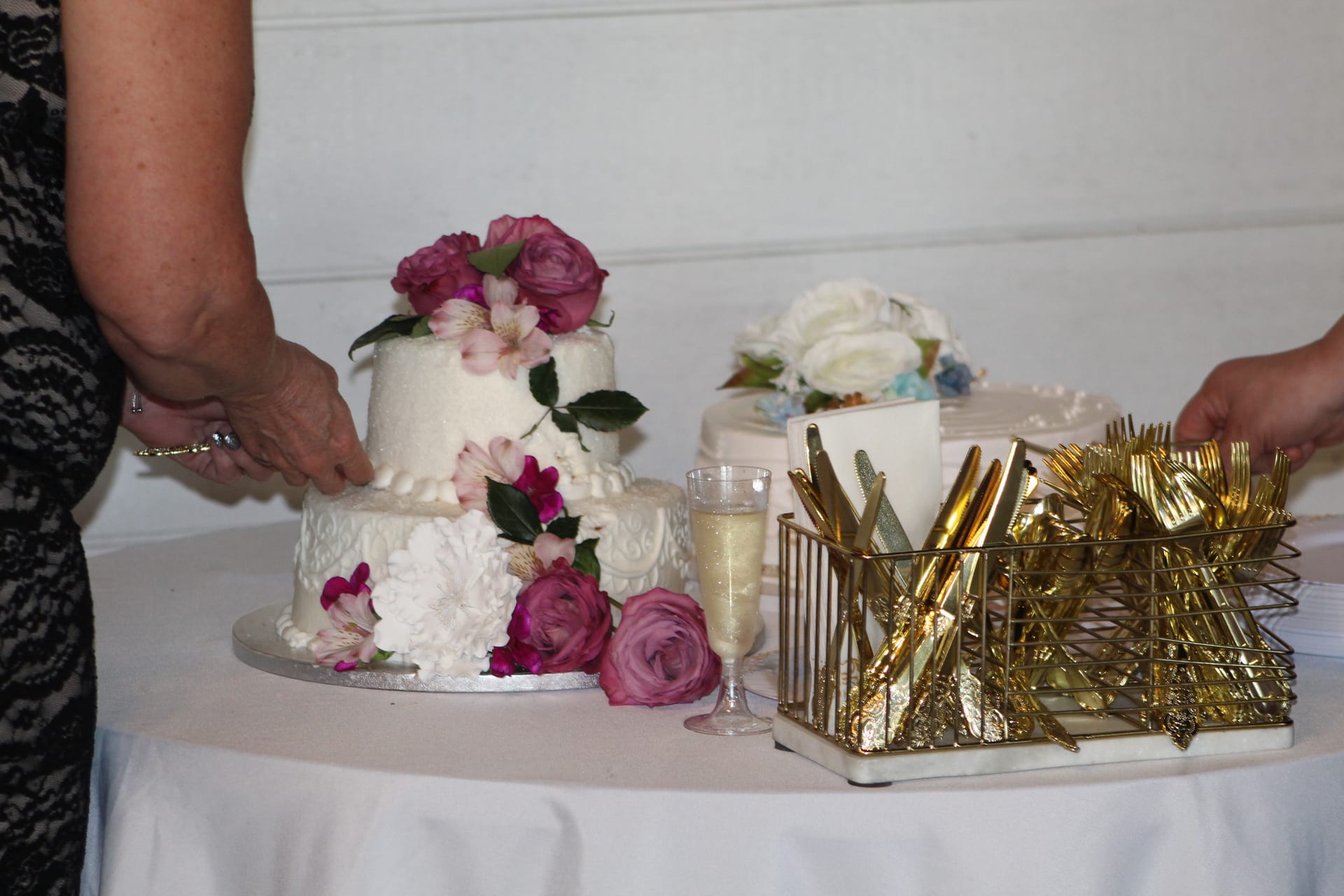 A two-tiered white wedding cake adorned with pink roses and alstroemeria flowers beside a gold cutlery caddy and champagne flute on a white-draped table at Scarborough House.