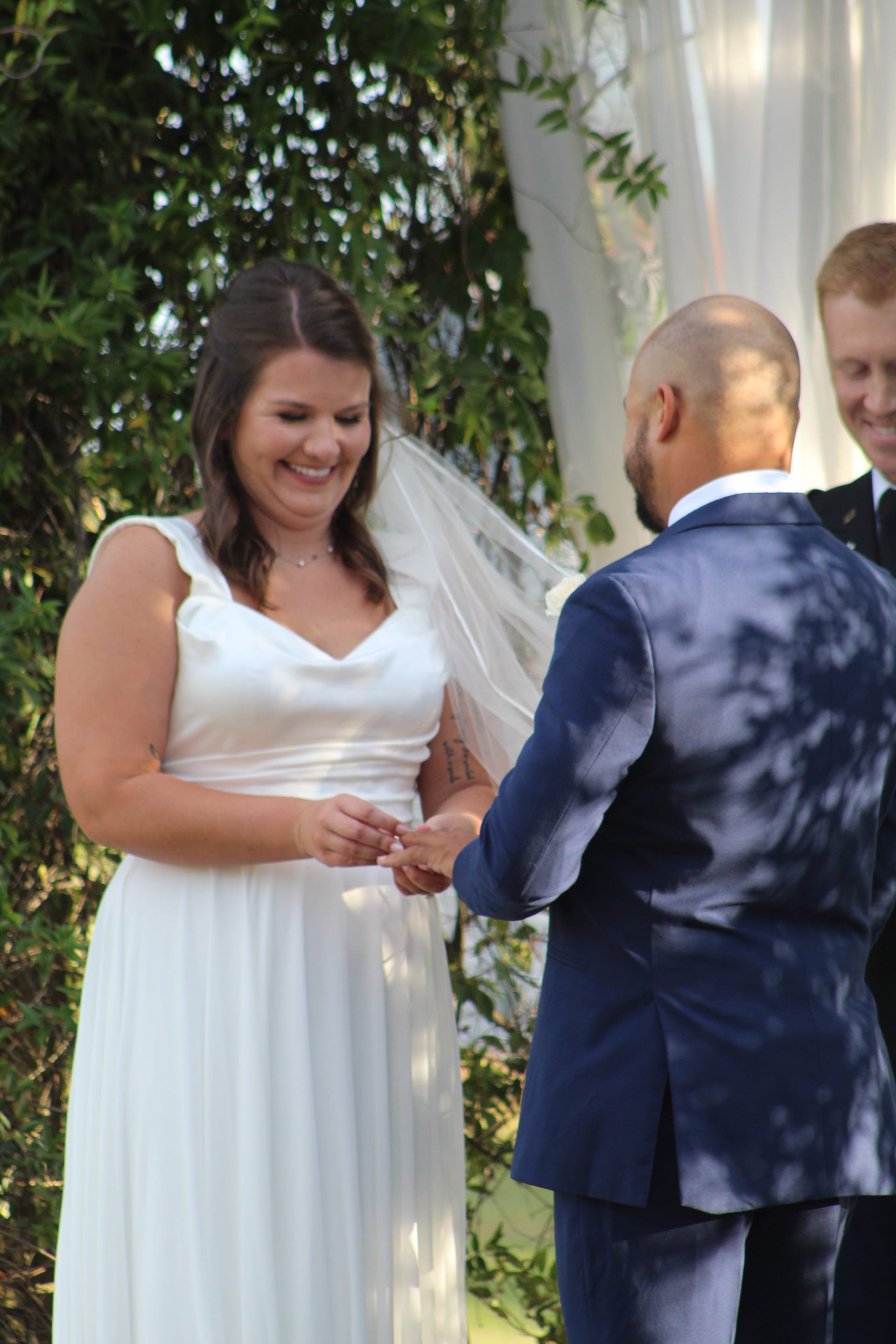 Smiling bride in a flowing white gown and veil exchanges rings with her groom in a navy suit during an outdoor wedding ceremony surrounded by lush greenery.