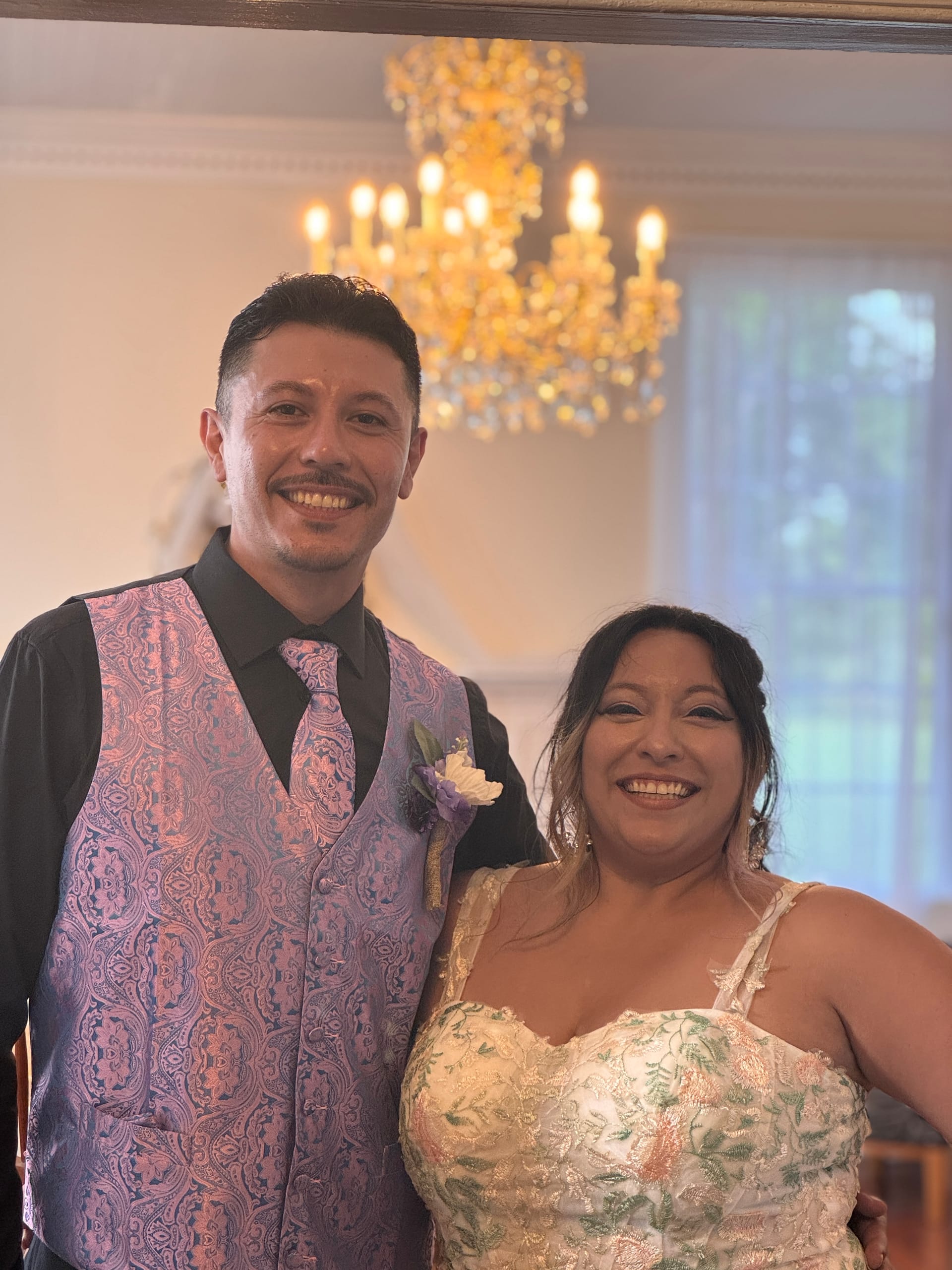 Smiling bride in a floral embroidered gown and groom in a paisley vest with boutonniere pose together beneath a golden chandelier inside the grand hall at Scarborough House.