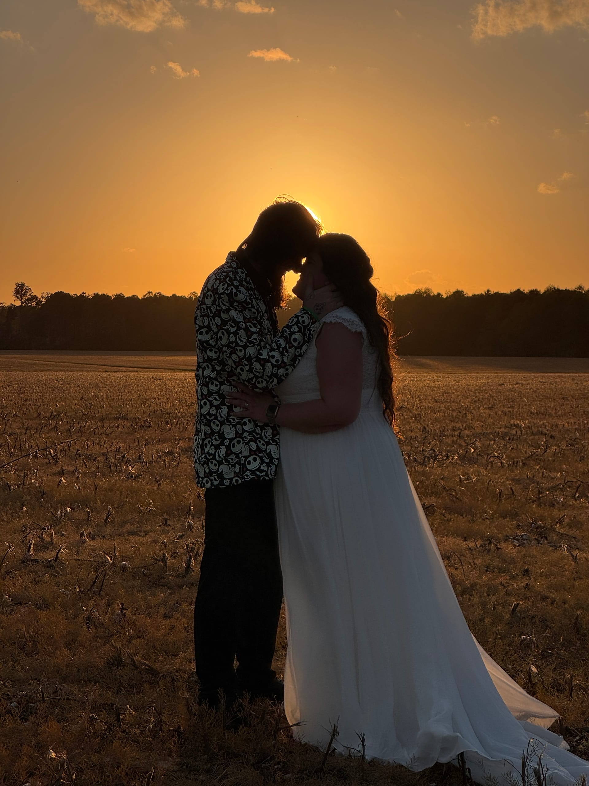 Silhouette of bride in flowing white gown and groom in patterned jacket touching foreheads tenderly against a vivid golden sunset over open farmland fields at Scarborough House.