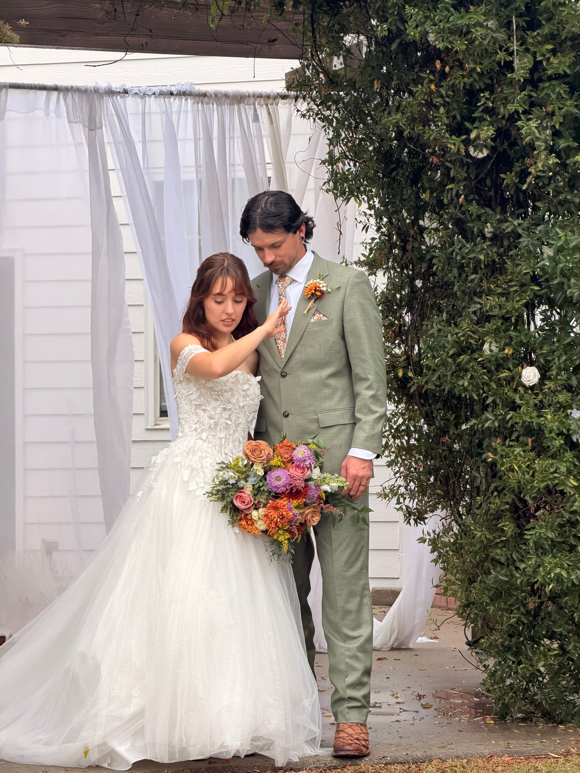 Bride in a floral appliqué ball gown adjusts her groom's floral tie as they stand together beneath white draped curtains beside a lush magnolia tree at Scarborough House estate.