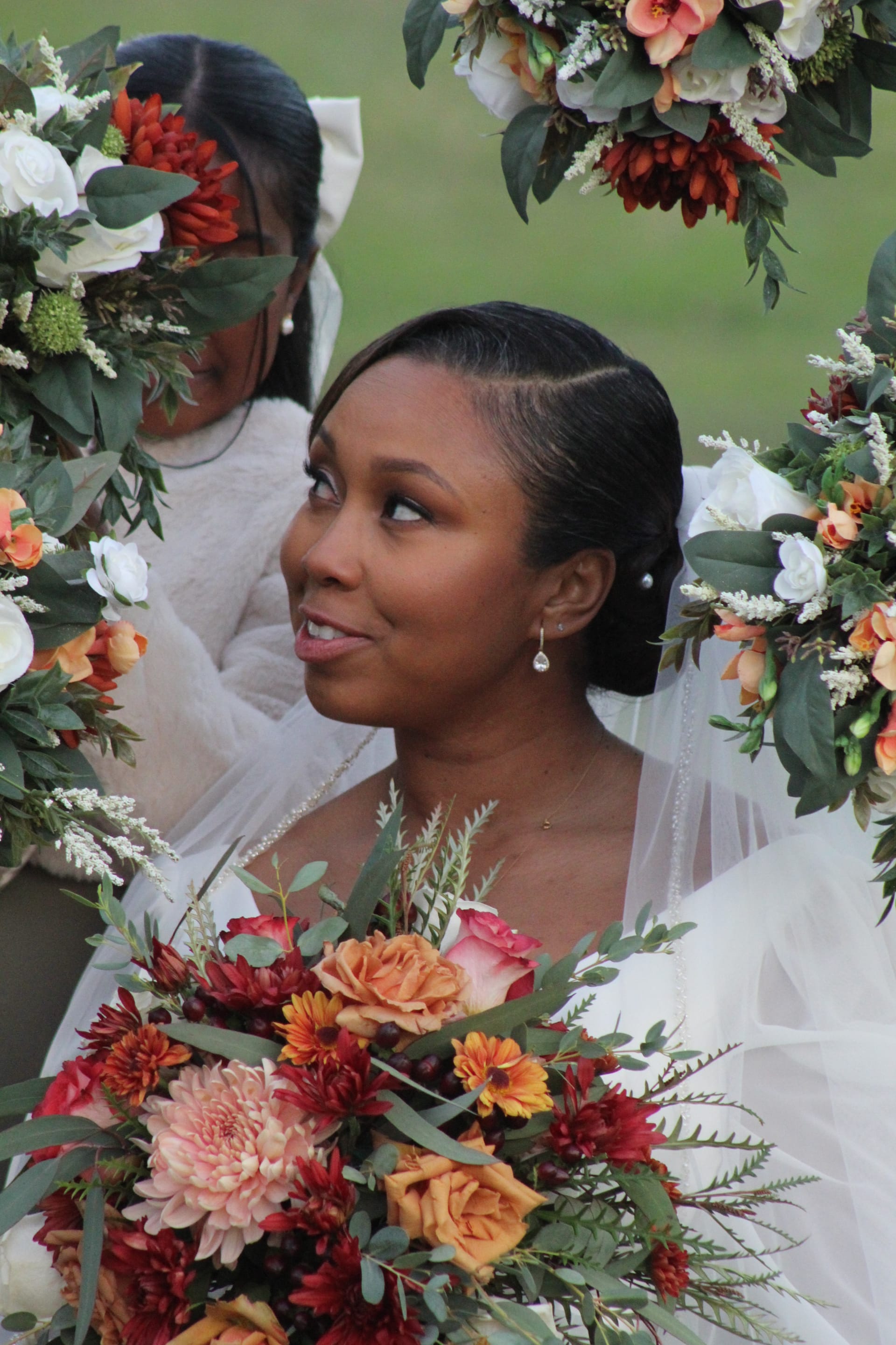 A bride in a white gown and veil gazes upward with a soft smile, framed by a lush autumn floral arch of burnt orange, burgundy, and blush blooms at Scarborough House estate.