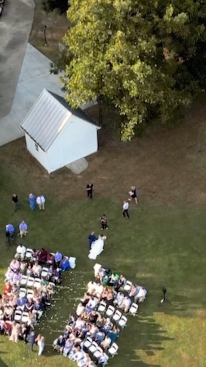 Aerial drone view of a bride in a white gown walking down a flower-petal aisle flanked by rows of seated guests on the sunlit ceremony lawn at Scarborough House estate.